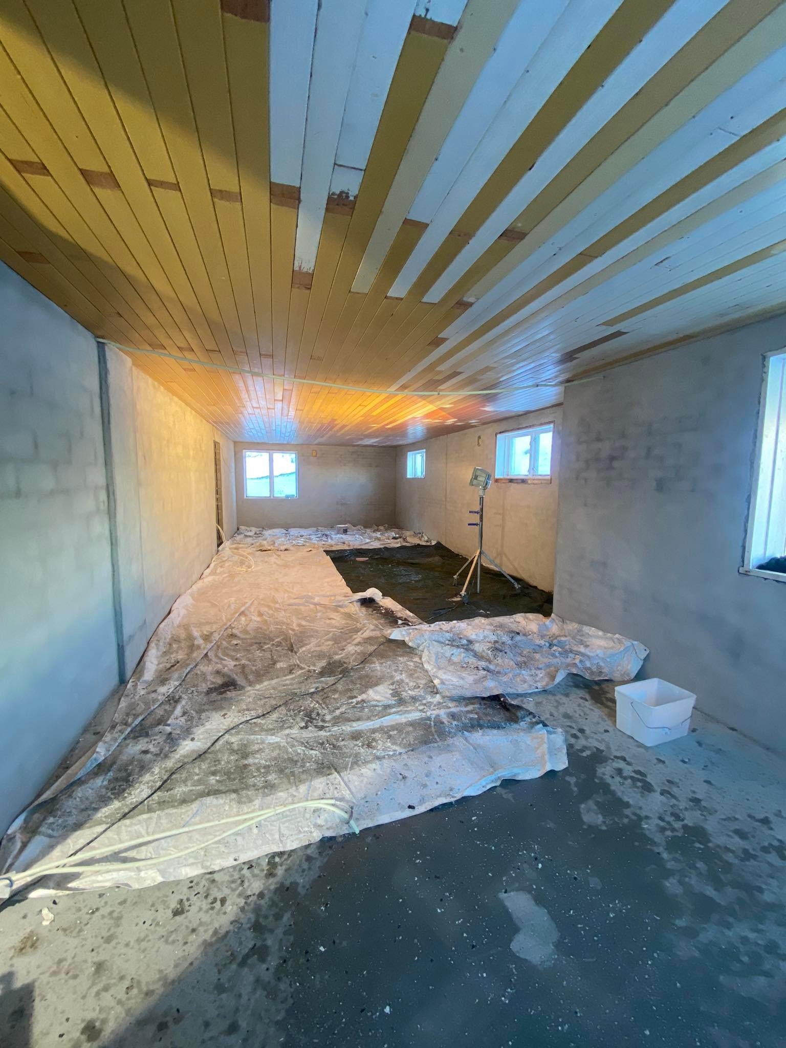 Wnętrze w trakcie prac przygotowawczych: zabezpieczona podłoga folią, ściany po wstępnym wyrównaniu, drewniany sufit częściowo pomalowany na biało, lampa budowlana na statywie.
