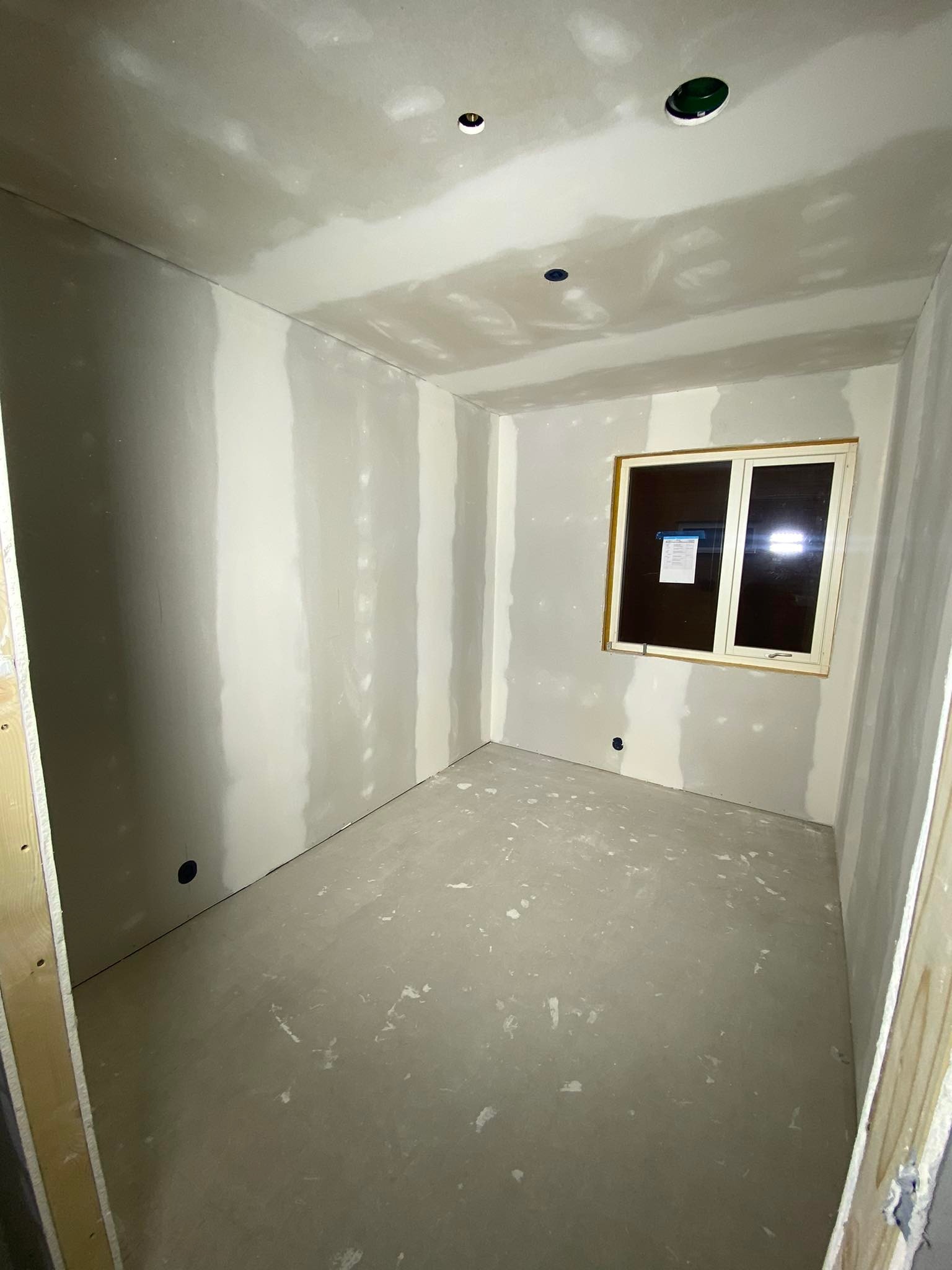 Surowe wnętrze pokoju z oknem po wstępnym nałożeniu gładzi szpachlowej na ściany i podłogę, widoczne otwory na punkty oświetleniowe w suficie.