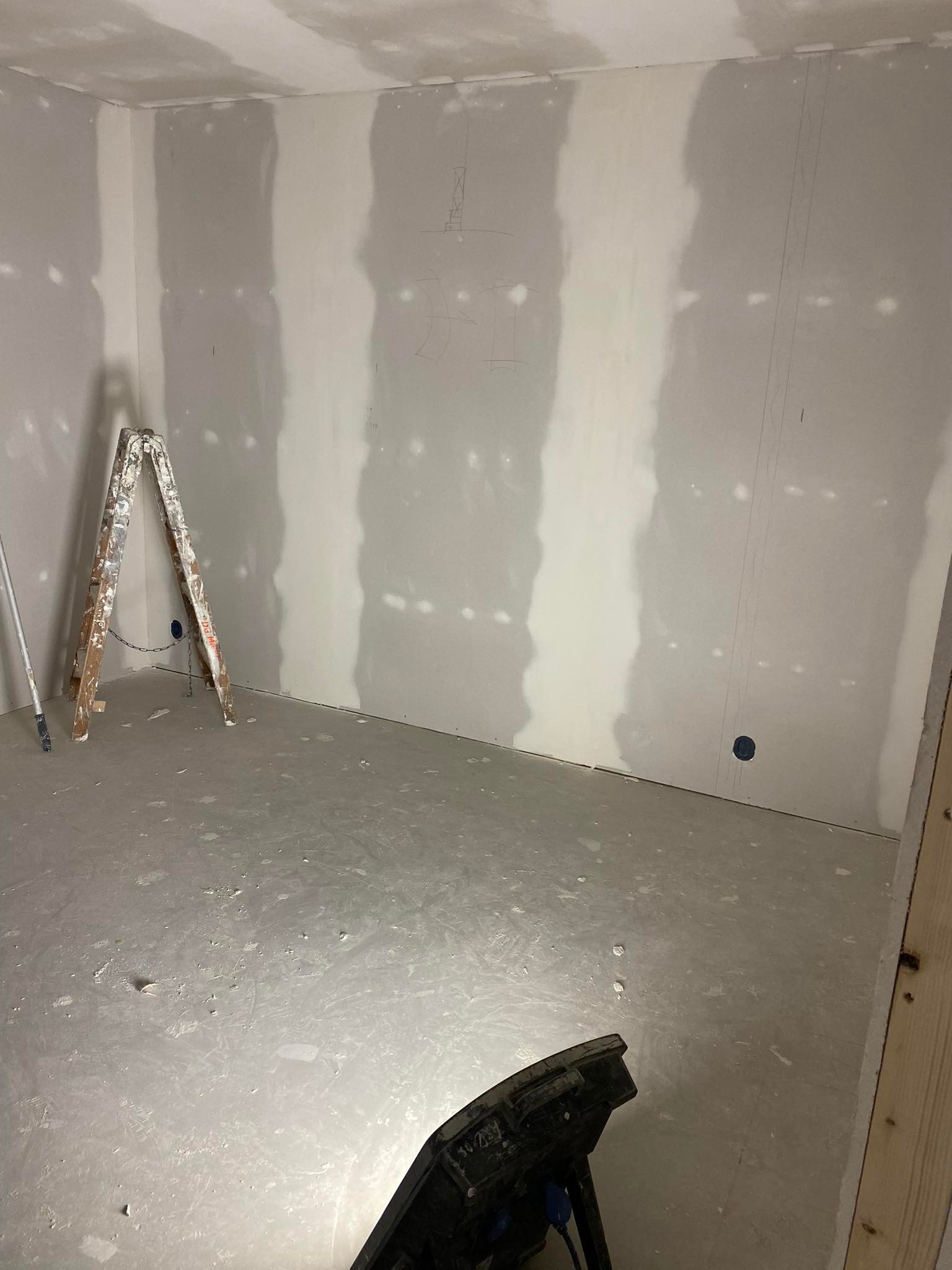 Pokój w trakcie prac przygotowawczych: ściany pokryte płytami gipsowo-kartonowymi, widoczne łączenia i ślady po szpachlowaniu, drabina malarska i lampa budowlana na podłodze.
