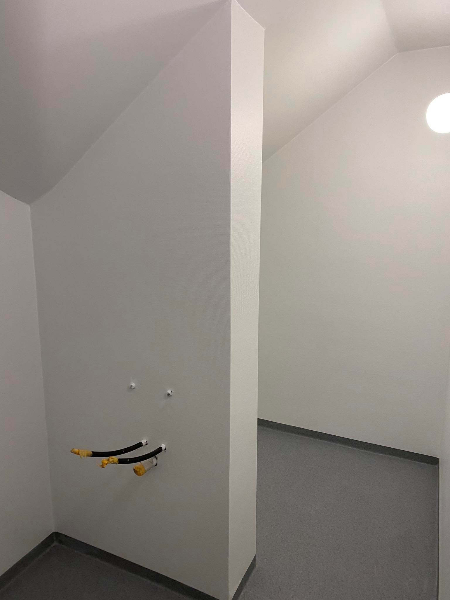 Wnętrze w stanie deweloperskim z widocznymi rurami instalacji wodnej wystającymi ze ściany, przygotowane do dalszych prac wykończeniowych, z szarą podłogą i białymi ścianami.