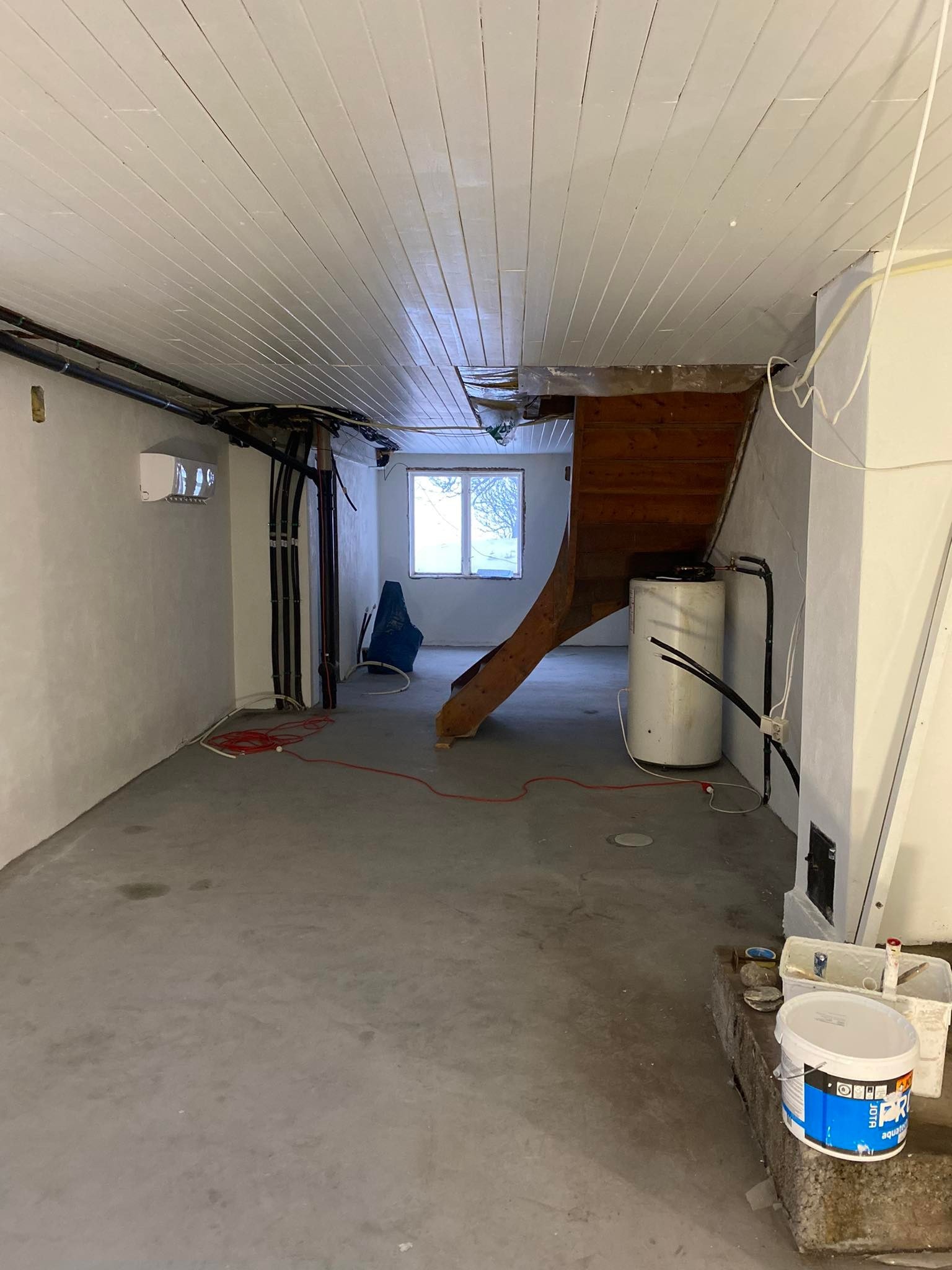 Piwnica w trakcie przygotowań do prac wykończeniowych: ściany po wstępnym wyrównaniu, widoczna instalacja elektryczna i hydrauliczna, drewniane schody z kotłem, pojemnik z farbą w rogu.