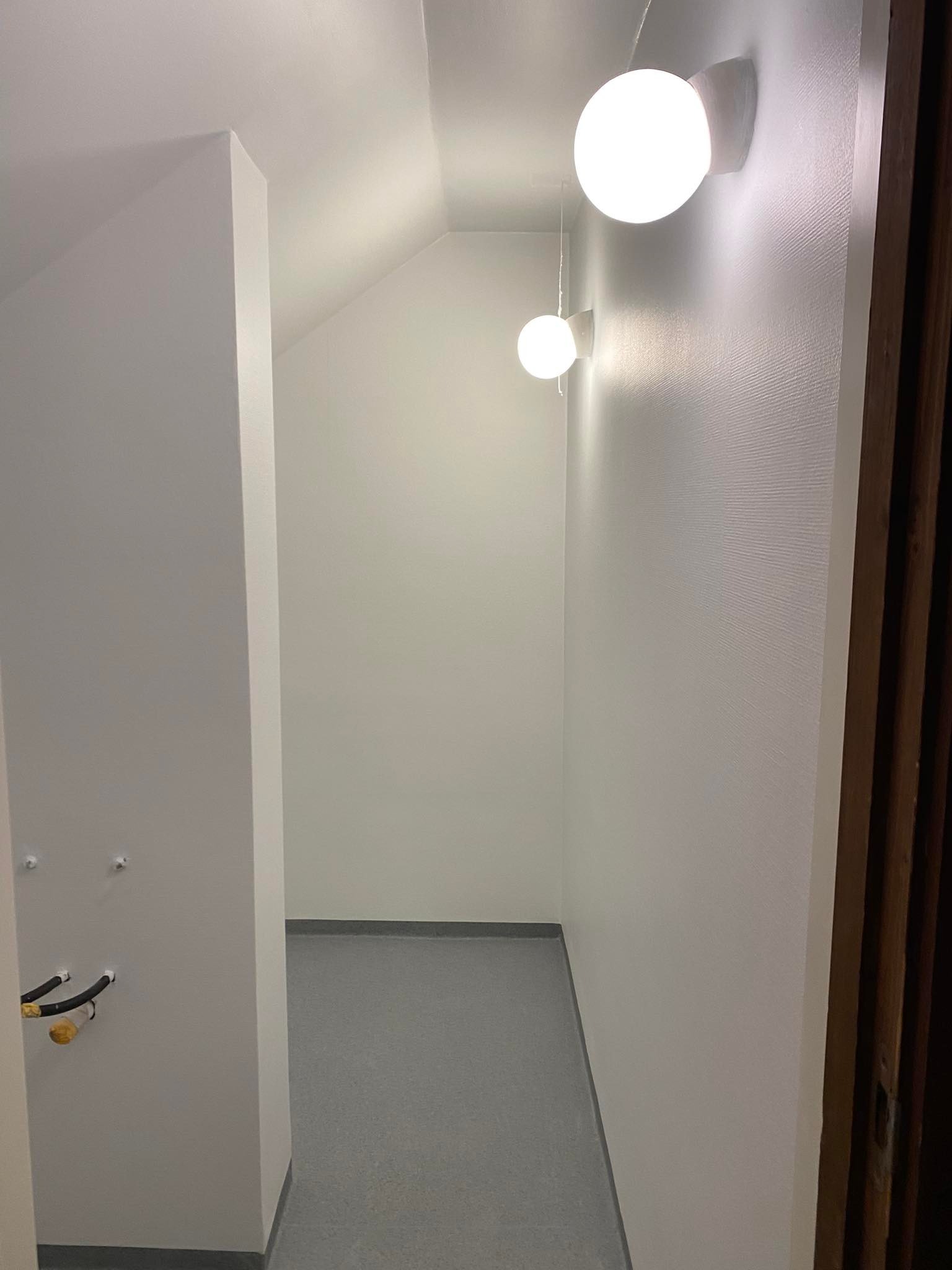 Wnętrze korytarza z szarą podłogą, białymi ścianami o delikatnej strukturze i dwoma okrągłymi lampami ściennymi emitującymi ciepłe światło, widoczne wystające rury instalacji.