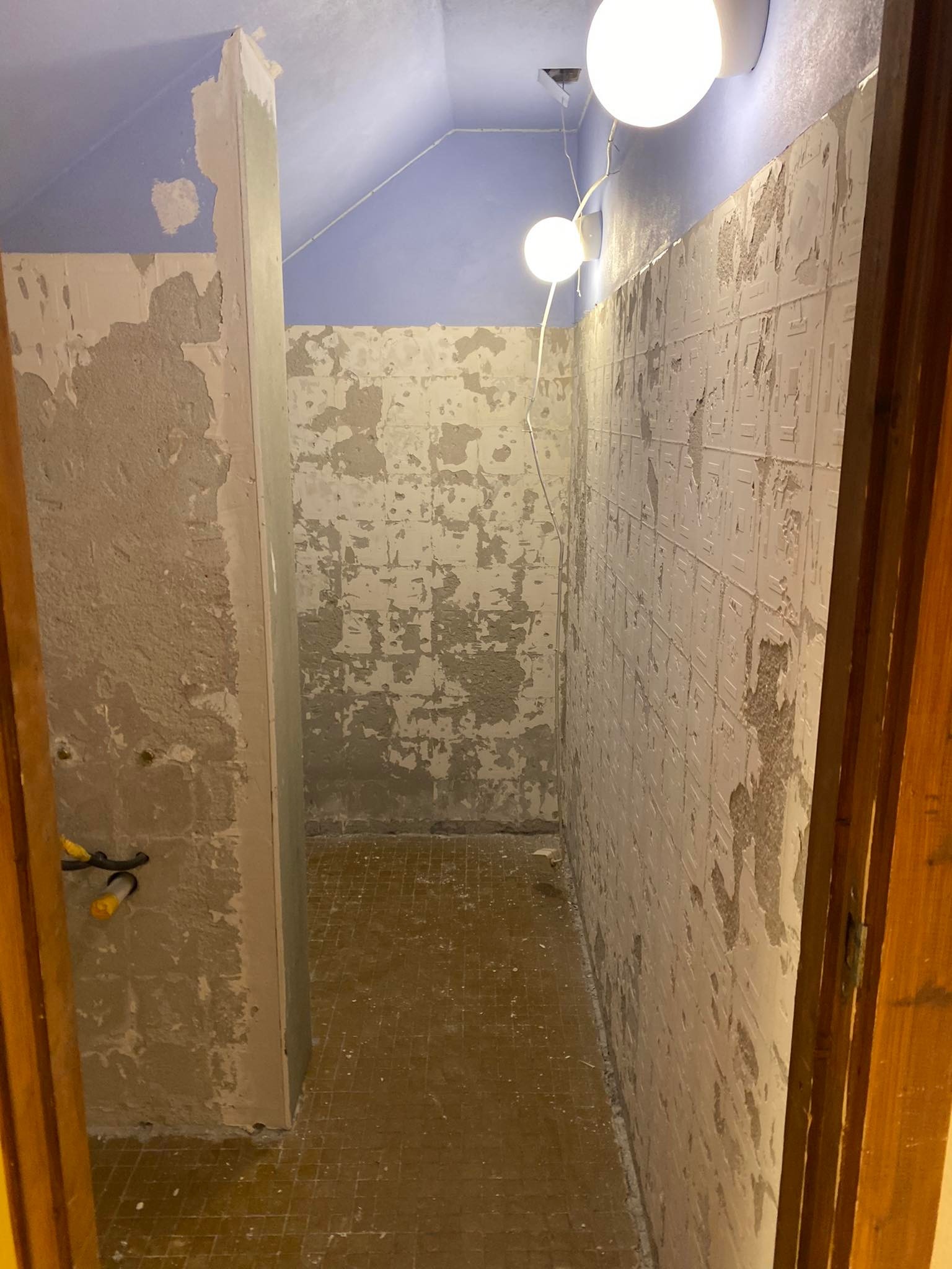 Łazienka w trakcie remontu: ściany częściowo pokryte resztkami starej glazury i szpachli, widoczne fragmenty instalacji elektrycznej, sufit pomalowany na niebiesko, żółtawa podłoga z małych...