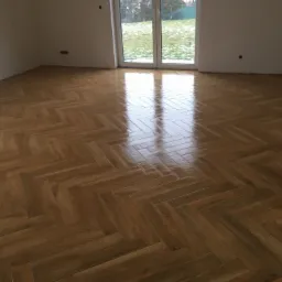 Ułożona, błyszcząca podłoga z drewnianych klepek w jodełkę w pustym pokoju z widokiem na ośnieżony ogród przez okno balkonowe.