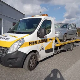 Biała laweta Renault z żółtymi akcentami i napisem 'Pomoc Drogowa' przewozi srebrny samochód osobowy na tle budynku i fragmentu ulicy.