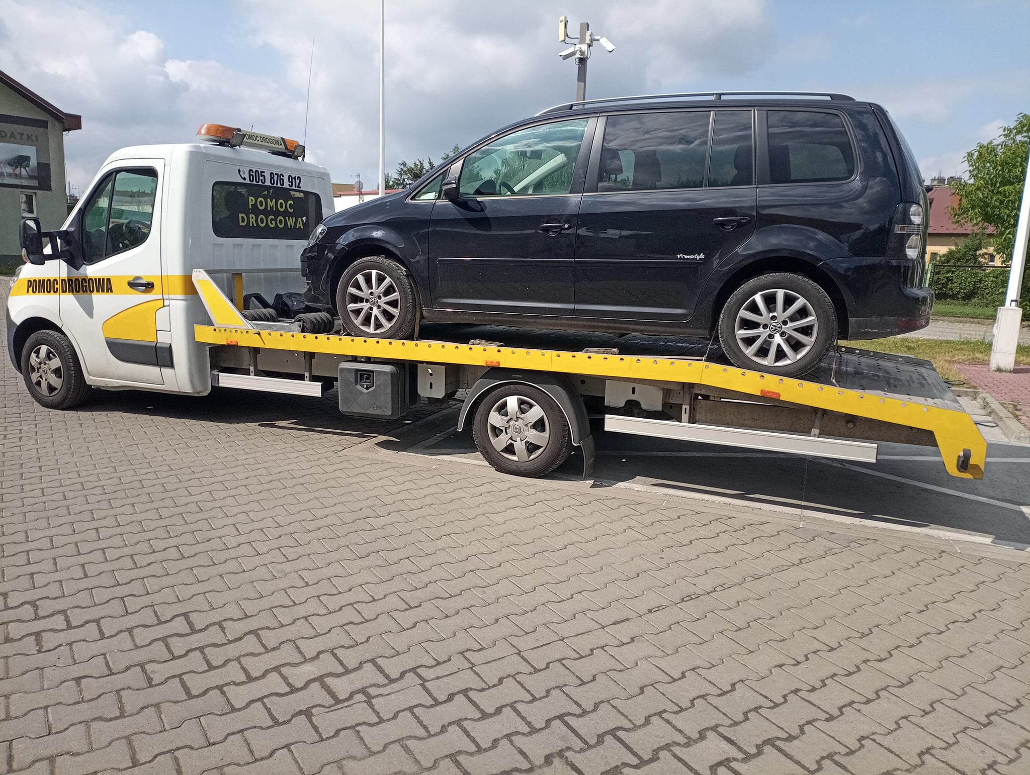 Czarny samochód osobowy typu van na żółtej lawecie z napisem 'Pomoc Drogowa' i numerem telefonu, zaparkowany na brukowanej powierzchni.