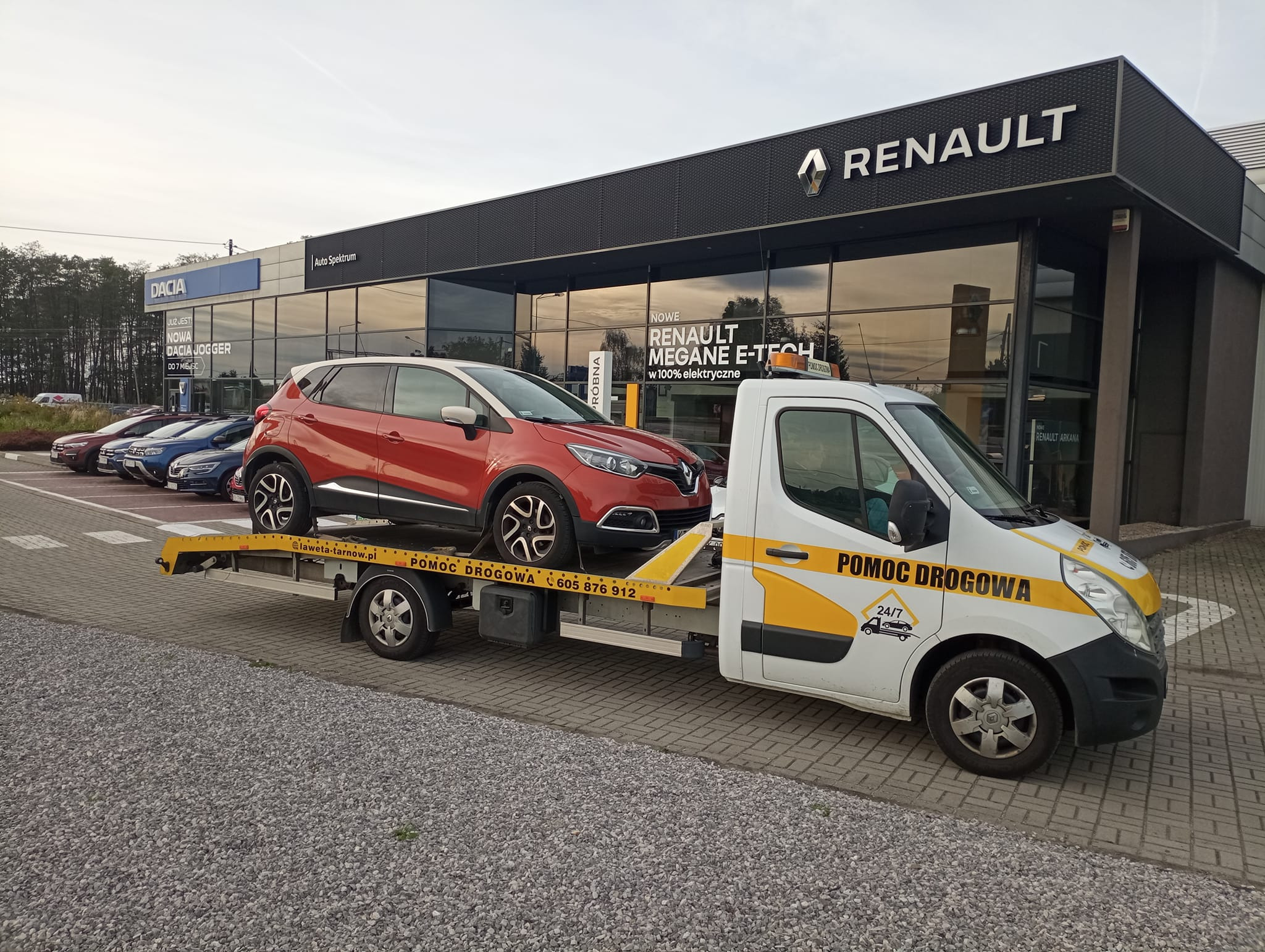 Pomarańczowy Renault Captur przewożony na żółtej lawecie na tle salonu samochodowego Renault i Dacia.