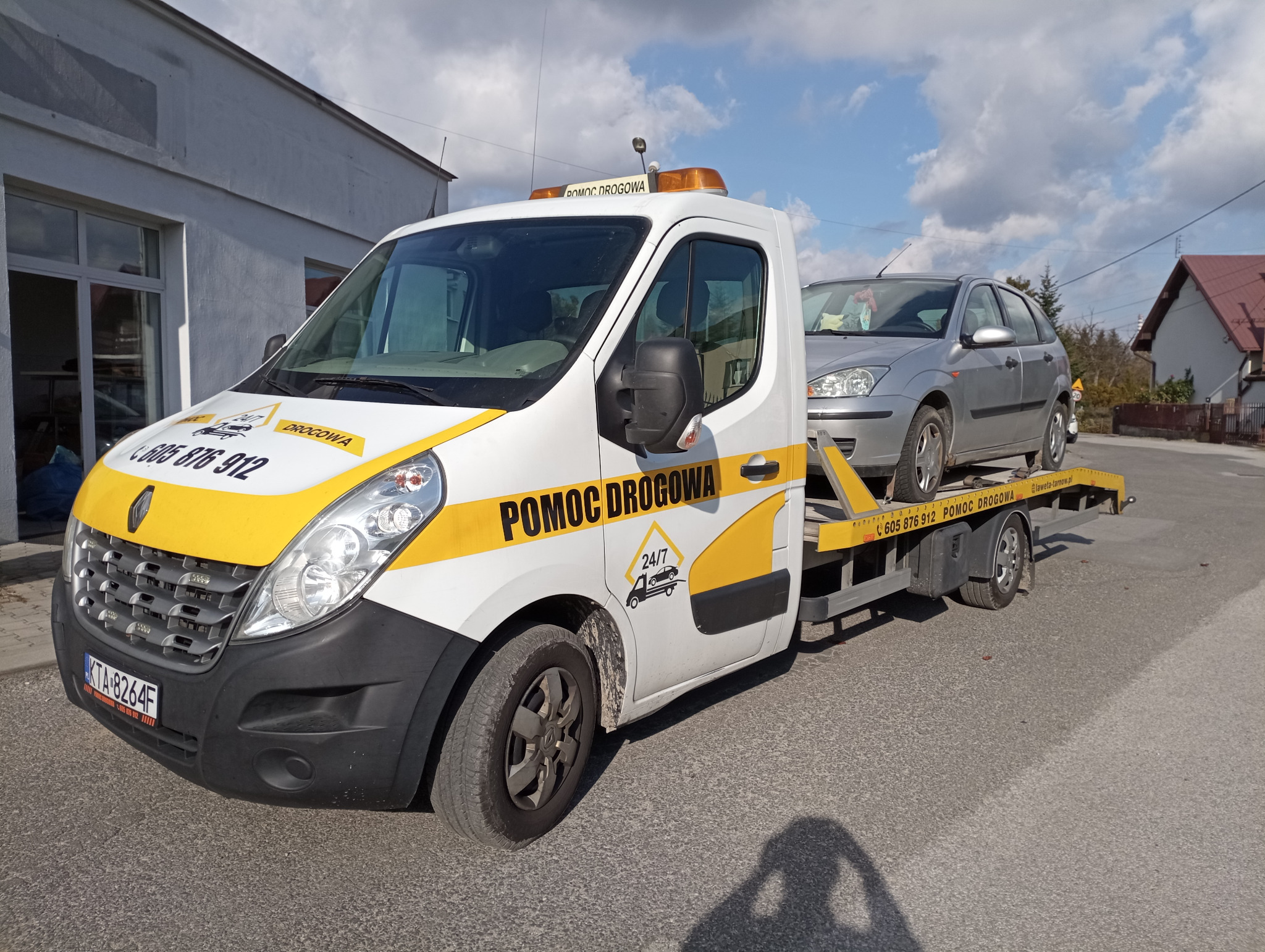 Biała laweta Renault z żółtymi akcentami i napisem 'Pomoc Drogowa' przewozi srebrny samochód osobowy na tle budynku i fragmentu ulicy.