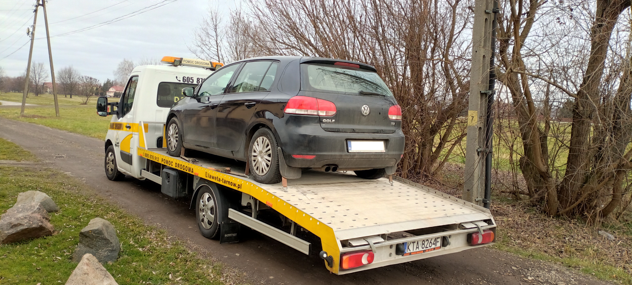 Czarny Volkswagen Golf na żółtej lawecie pomocy drogowej na polnej drodze, widoczne tablice rejestracyjne lawety i numer telefonu pomocy drogowej na kabinie.