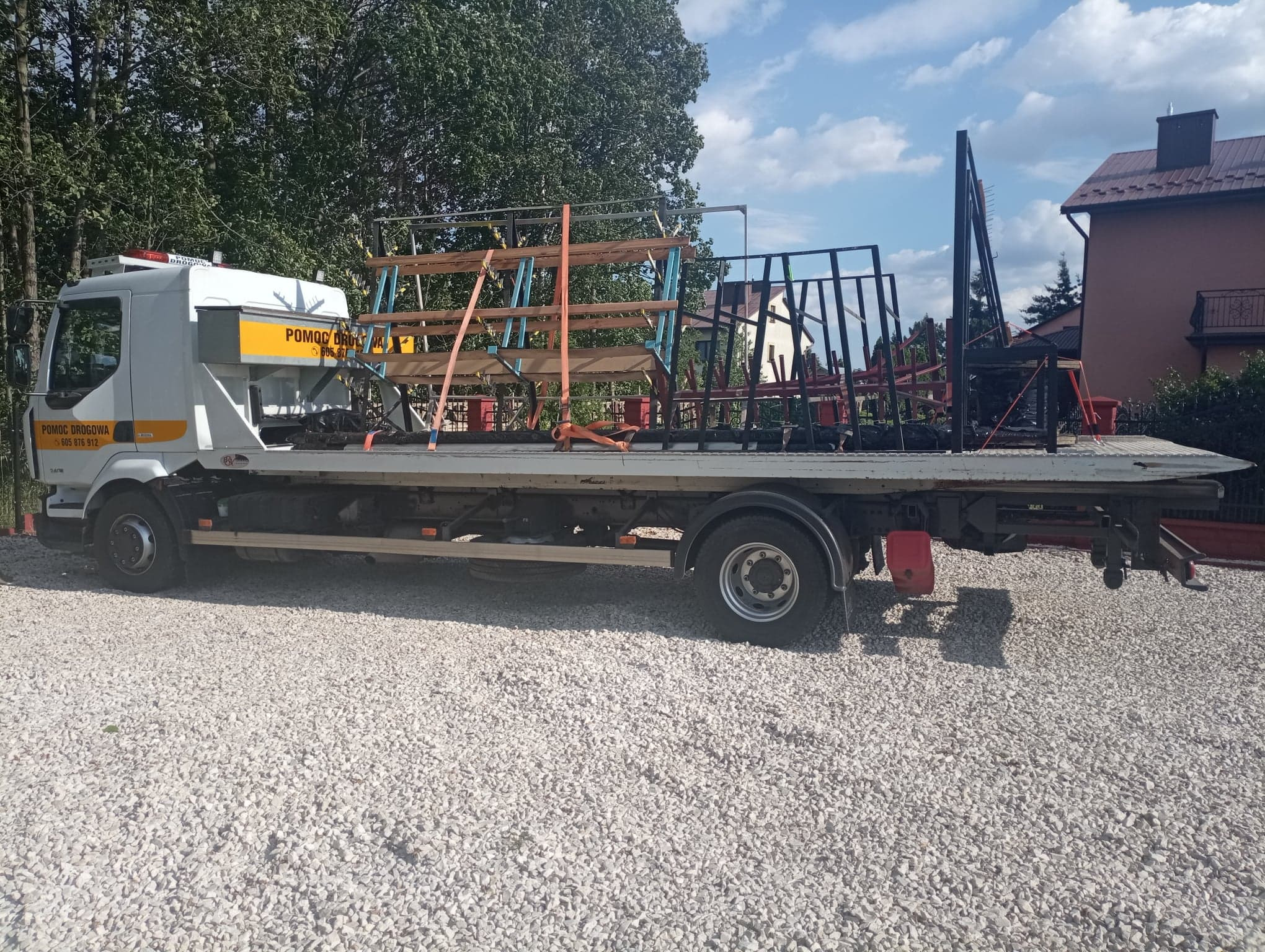 Laweta z konstrukcją do przewozu okien lub elementów szklanych, zaparkowana na żwirowym podłożu, budynek mieszkalny i drzewa w tle.