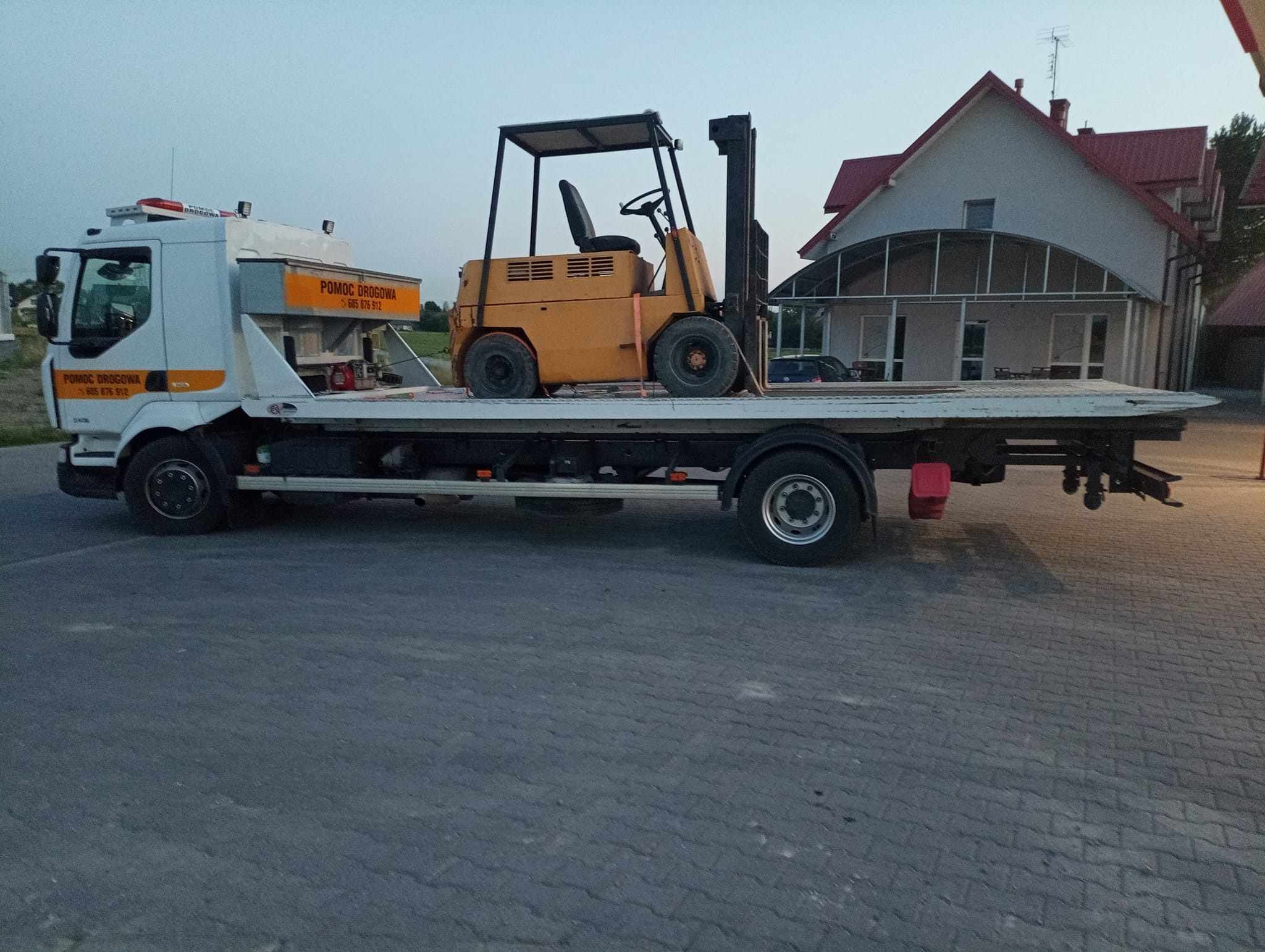 Żółty wózek widłowy zabezpieczony pasami na platformie lawety z napisem 'Pomoc Drogowa' na tle budynku mieszkalnego.