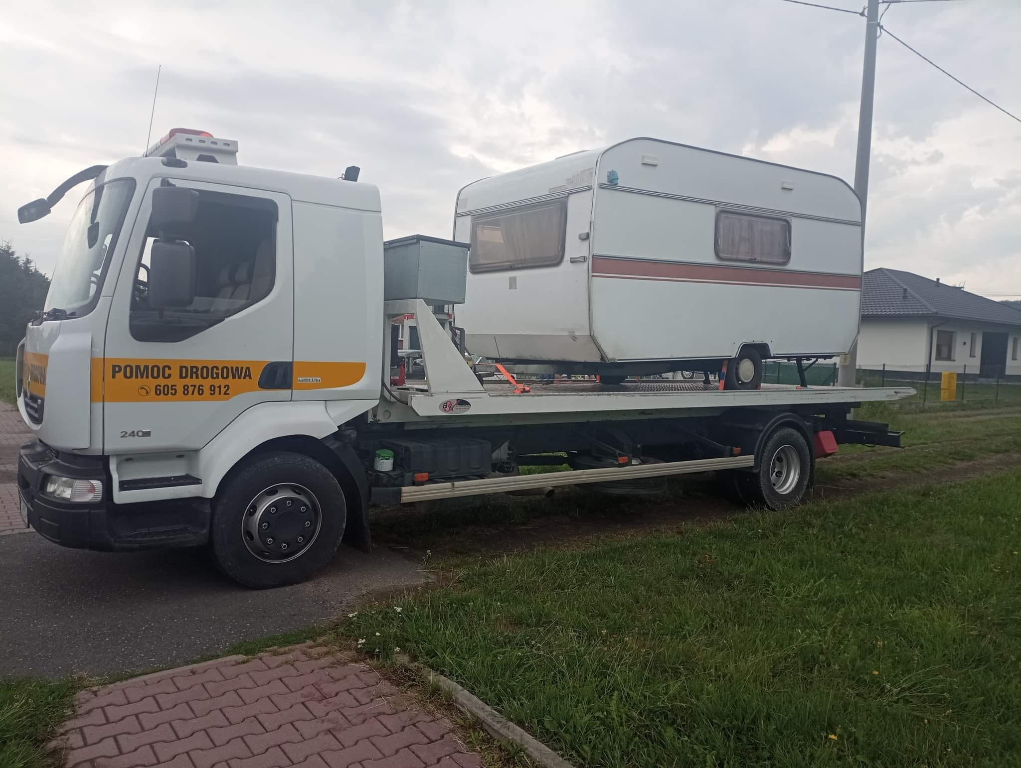 Biała laweta z napisem 'Pomoc Drogowa' przewozi białą przyczepę kempingową z brązowym pasem na tle pochmurnego nieba i domów.