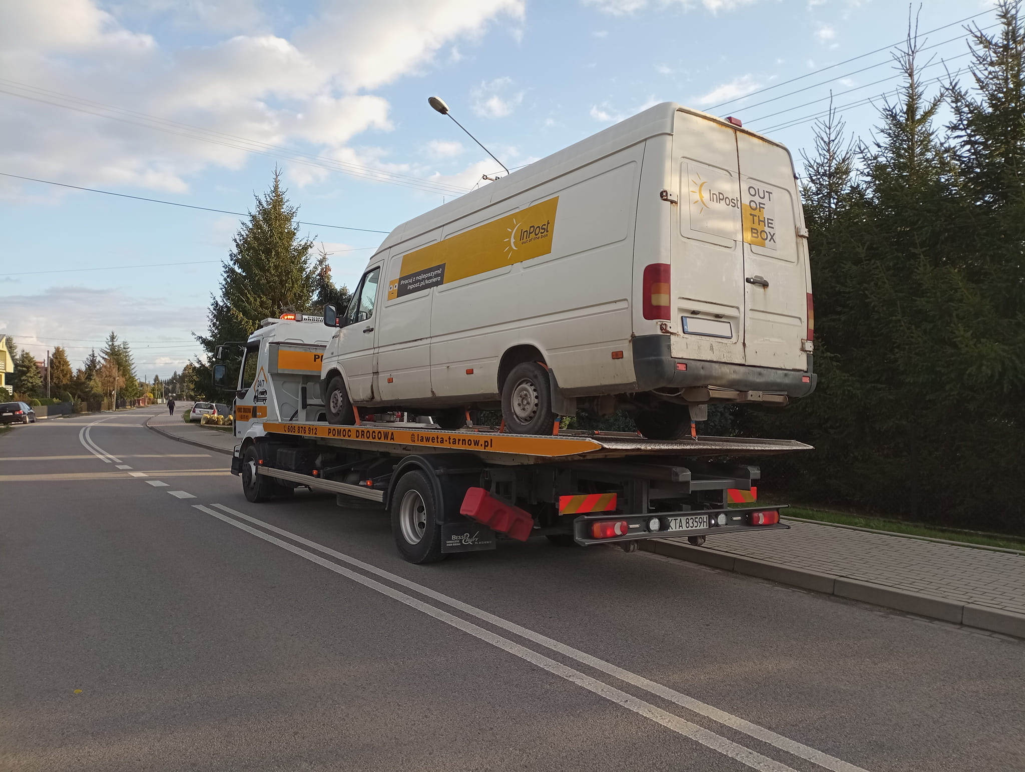 Laweta z białym dostawczym busem InPost na platformie, widok z tyłu na ulicy w słoneczny dzień.