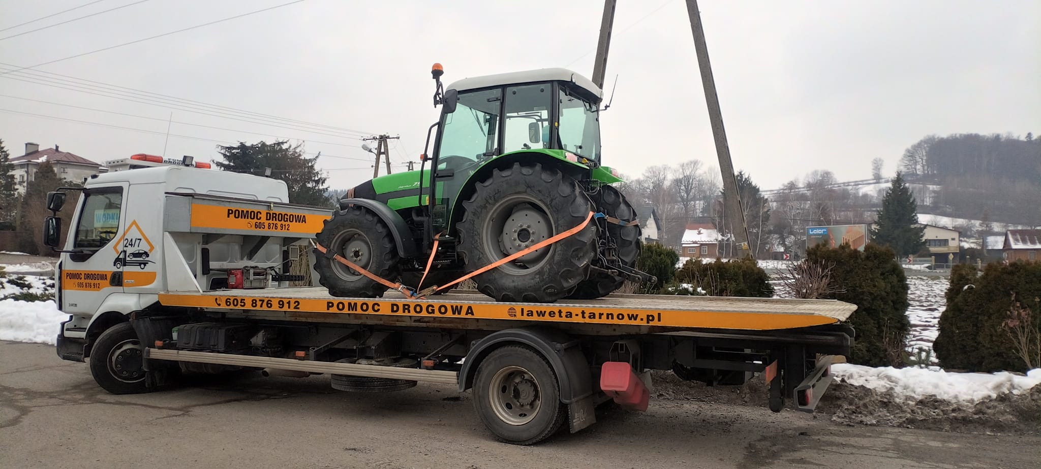 Zielony traktor zabezpieczony pomarańczowymi pasami na platformie lawety pomocy drogowej z napisem 'POMOC DROGOWA 605 876 912' i adresem laweta-tarnow.pl w zimowej scenerii.