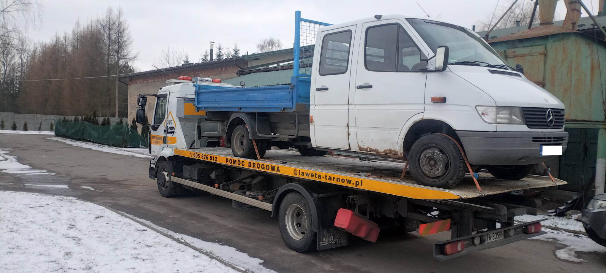 Biały dostawczy samochód z rdzawymi plamami na karoserii, zabezpieczony pasami, przewożony na żółtej lawecie z niebieską platformą, na tle zimowego krajobrazu z budynkami i drzewami.