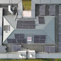 Widok z góry na szary dach pokryty panelami fotowoltaicznymi różnej wielkości, z dodatkowym agregatem chłodniczym i świetlikiem dachowym.