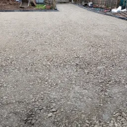 Podłoże z kruszywa przygotowane pod budowę chodnika lub podjazdu, widoczny brzeg z obrzeżami, w tle budynek i maszyna budowlana.