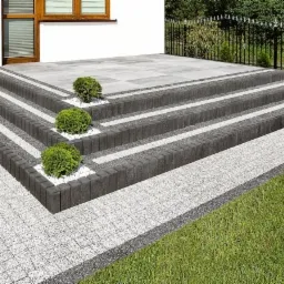 Schody zewnętrzne wyłożone szarą kostką brukową z ciemnymi obrzeżami, ozdobione białym grysem i kulistymi krzewami, prowadzące do domu z oknem i metalowym ogrodzeniem w tle.