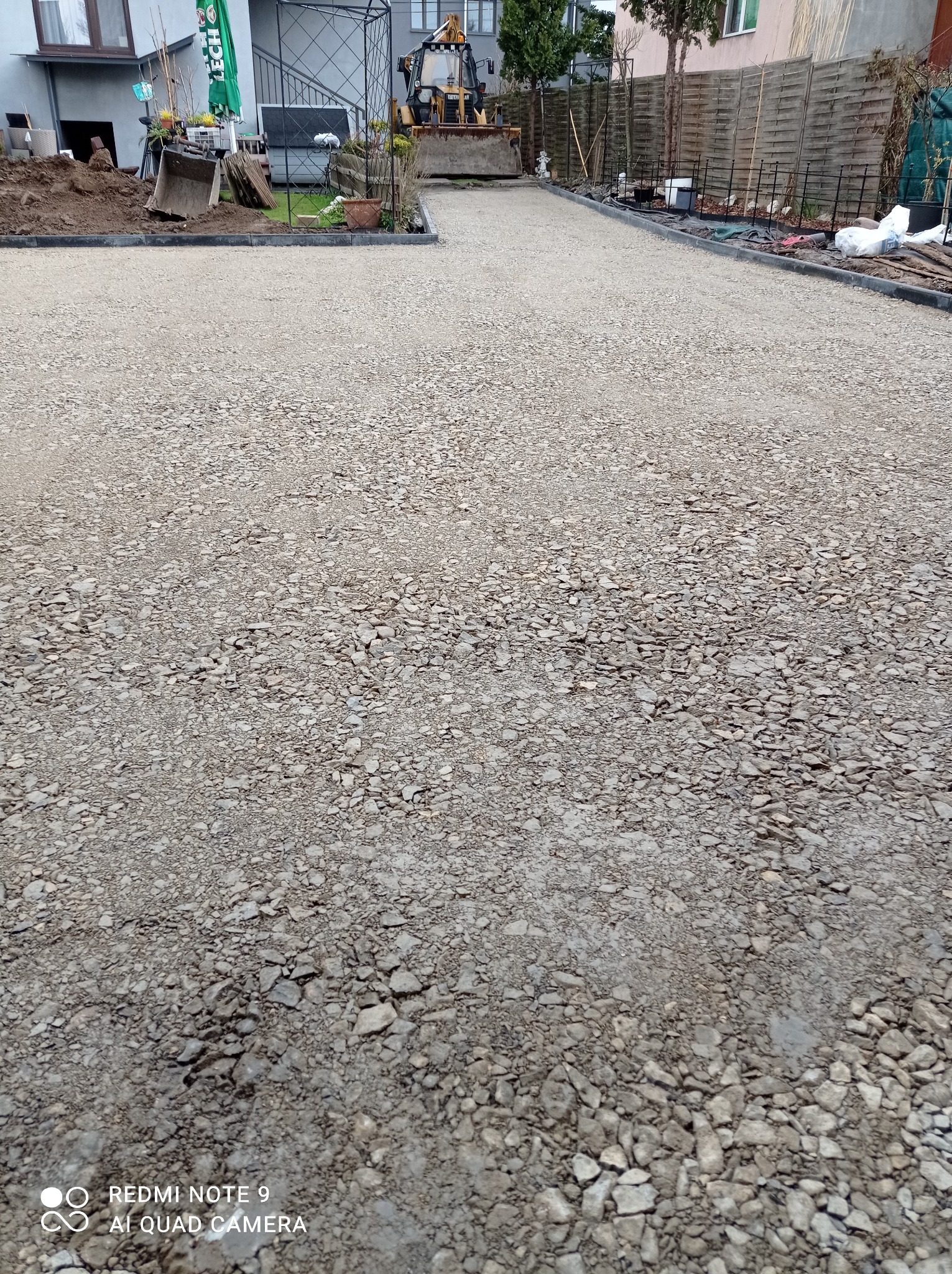 Podłoże z kruszywa przygotowane pod budowę chodnika lub podjazdu, widoczny brzeg z obrzeżami, w tle budynek i maszyna budowlana.