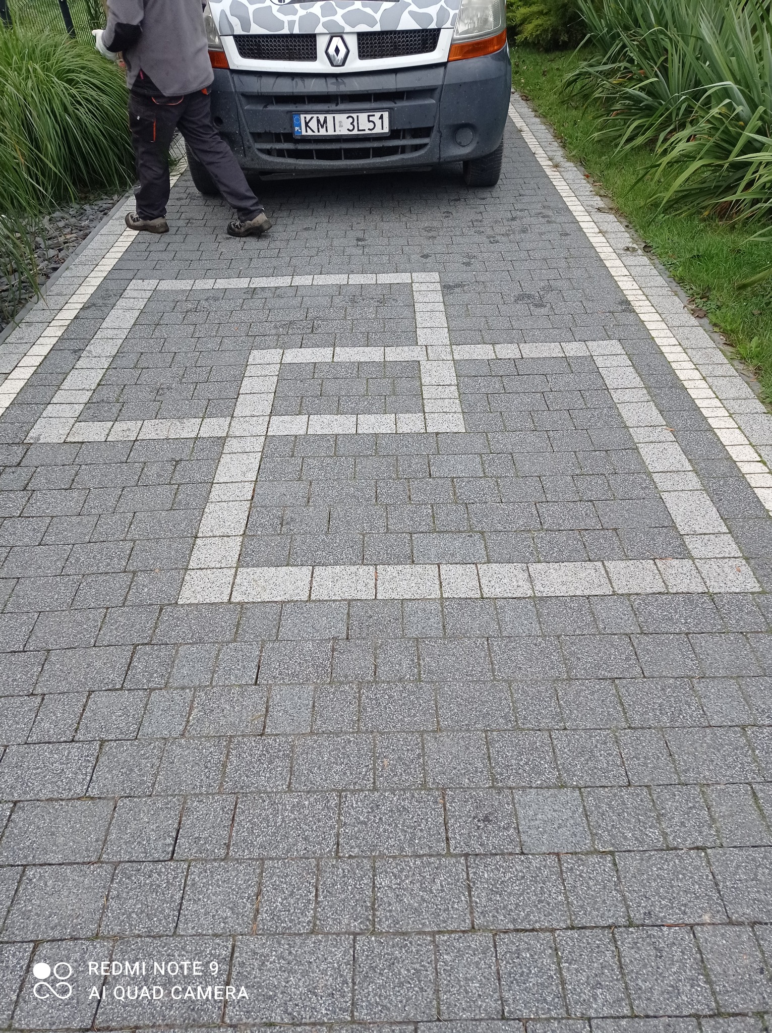 Szara kostka brukowa z białym wzorem geometrycznym, częściowo zasłonięta przez zaparkowany samochód dostawczy Renault i osobę w odzieży roboczej, z zielenią w tle.