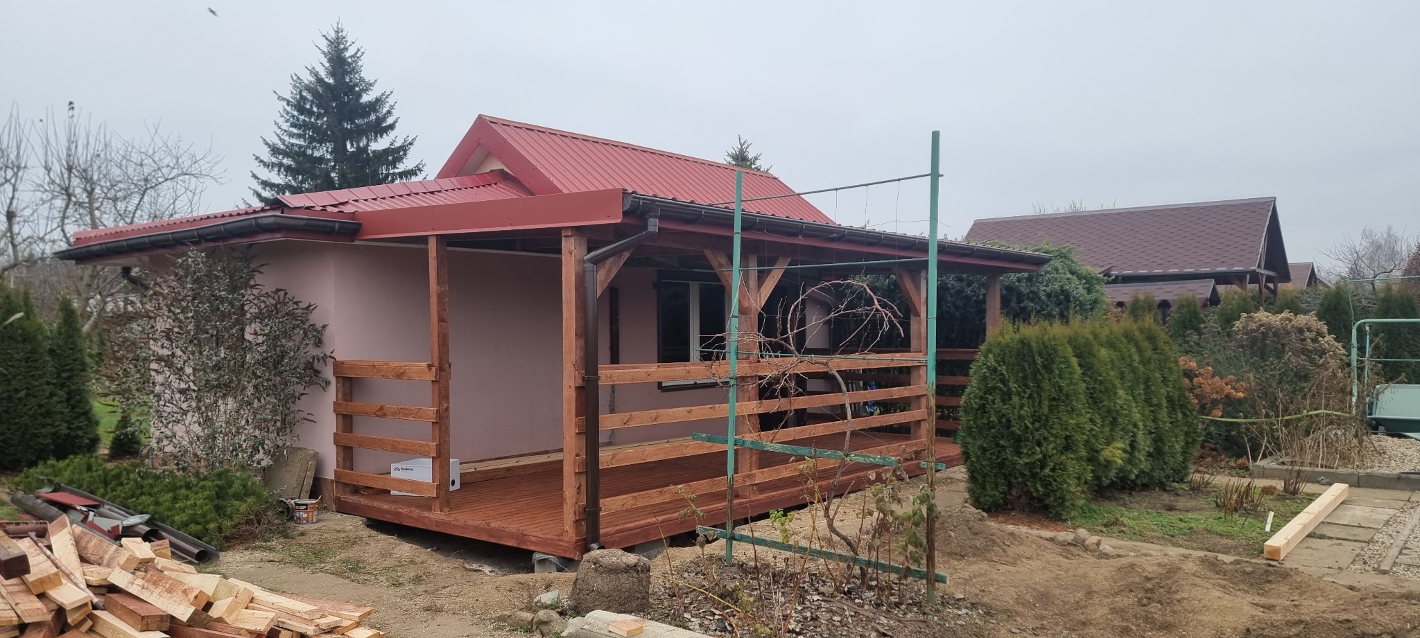 Nowo wybudowana drewniana altana z czerwonym dachem blachodachówką, widoczne świeżo pomalowane drewniane elementy, fragment budynku mieszkalnego i przygotowane drewno do dalszych prac.