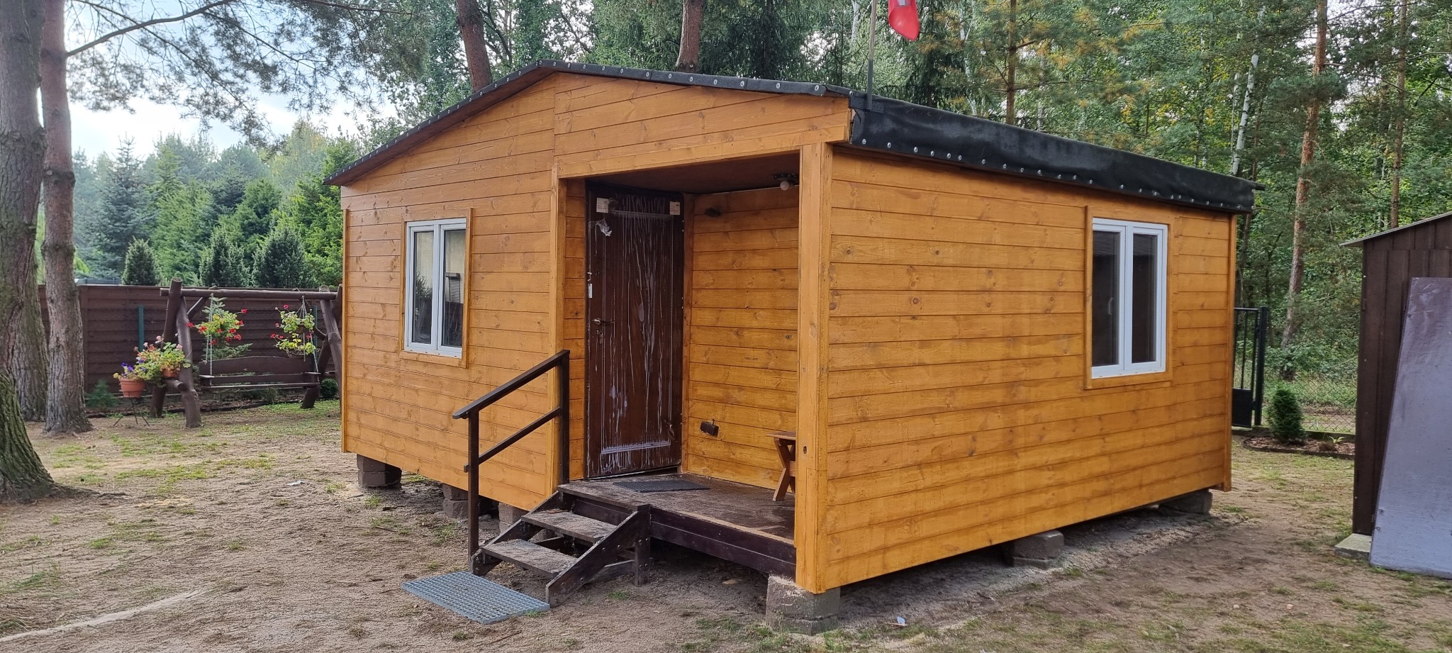Drewniany domek letniskowy z oknami i schodami wejściowymi, otoczony drzewami i zielenią, widoczny w dziennym świetle.