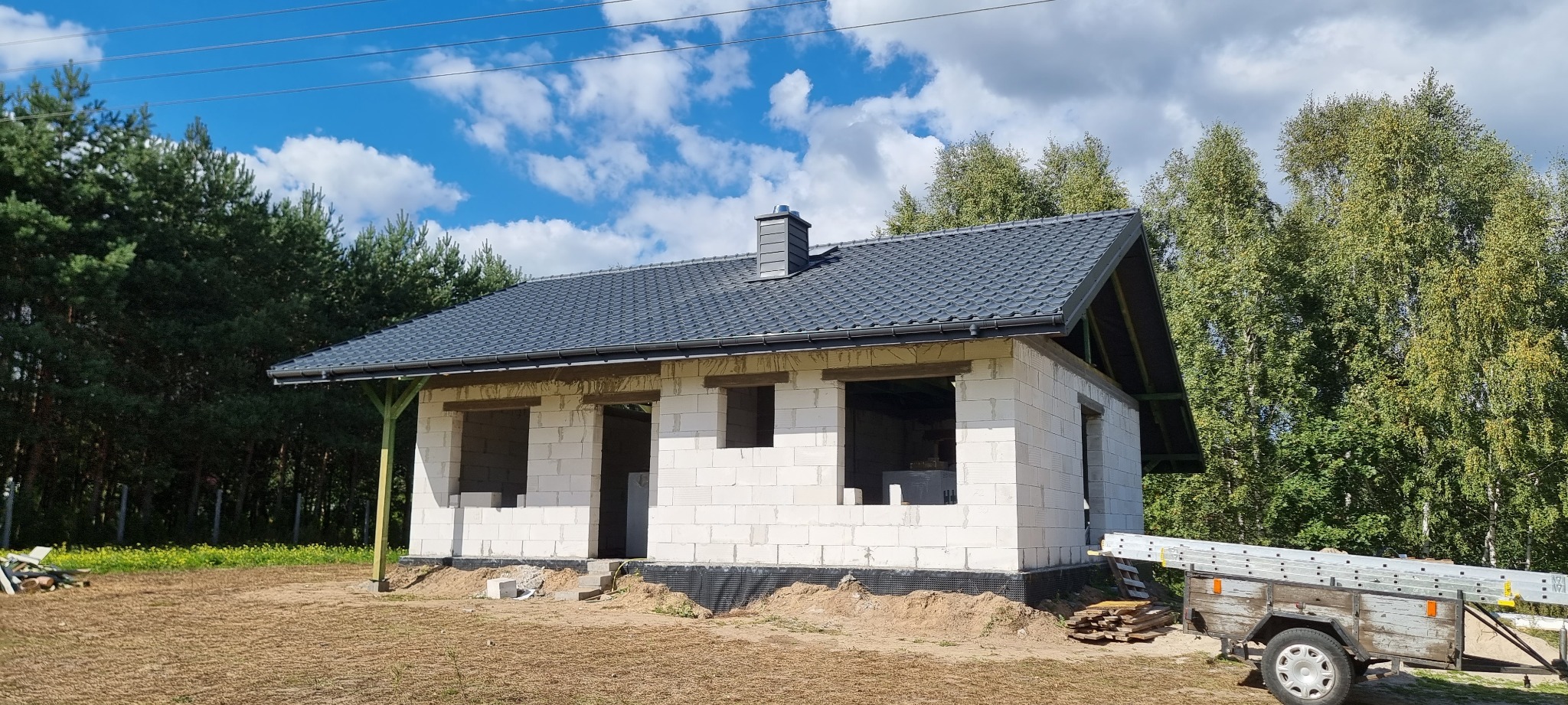 Surowy budynek z białych bloczków z ukończonym, ciemnoszarym dachem z kominem na tle błękitnego nieba i drzew, przyczepka z drabiną obok.