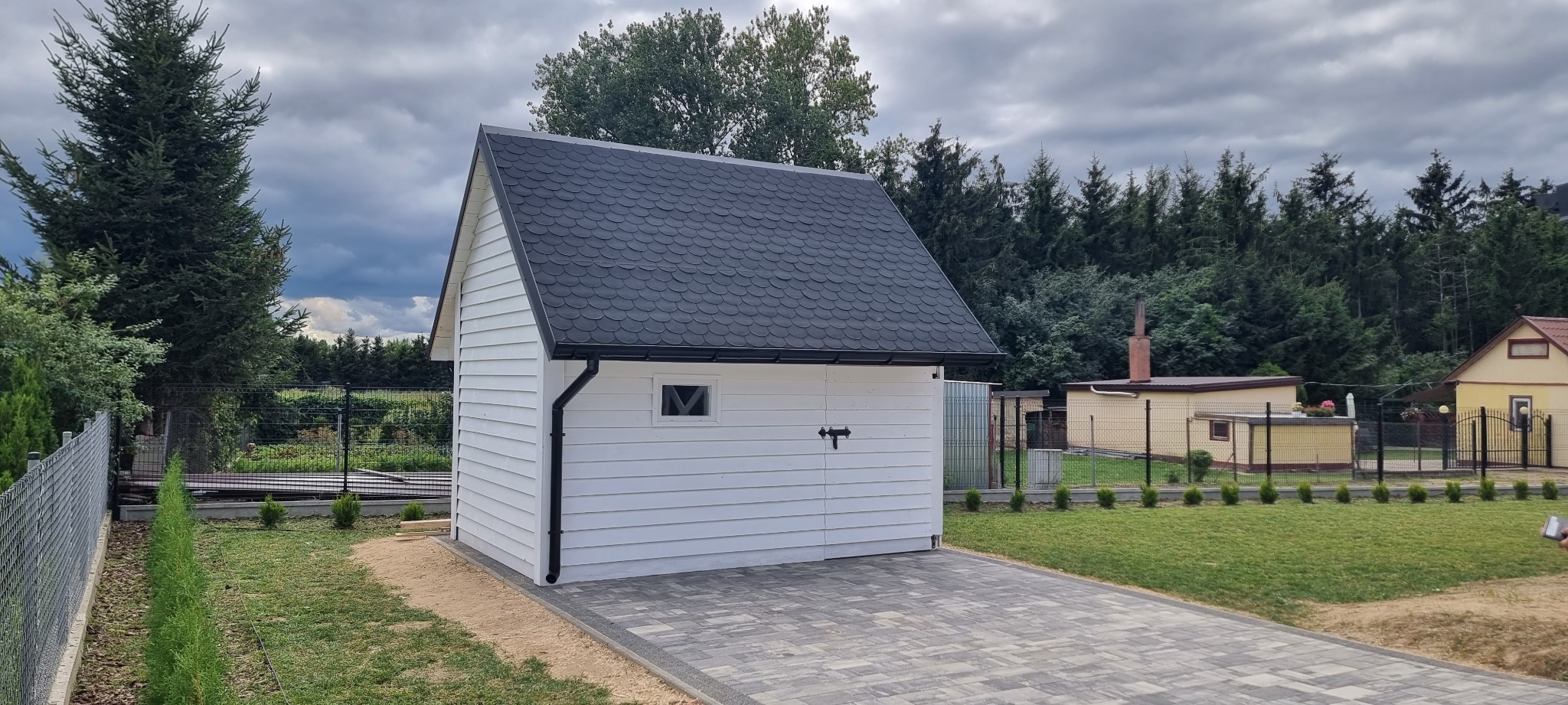 Nowoczesny biały domek narzędziowy z czarnym dachem z gontów bitumicznych, widok z zewnątrz, otoczony zielonym trawnikiem i drzewami.