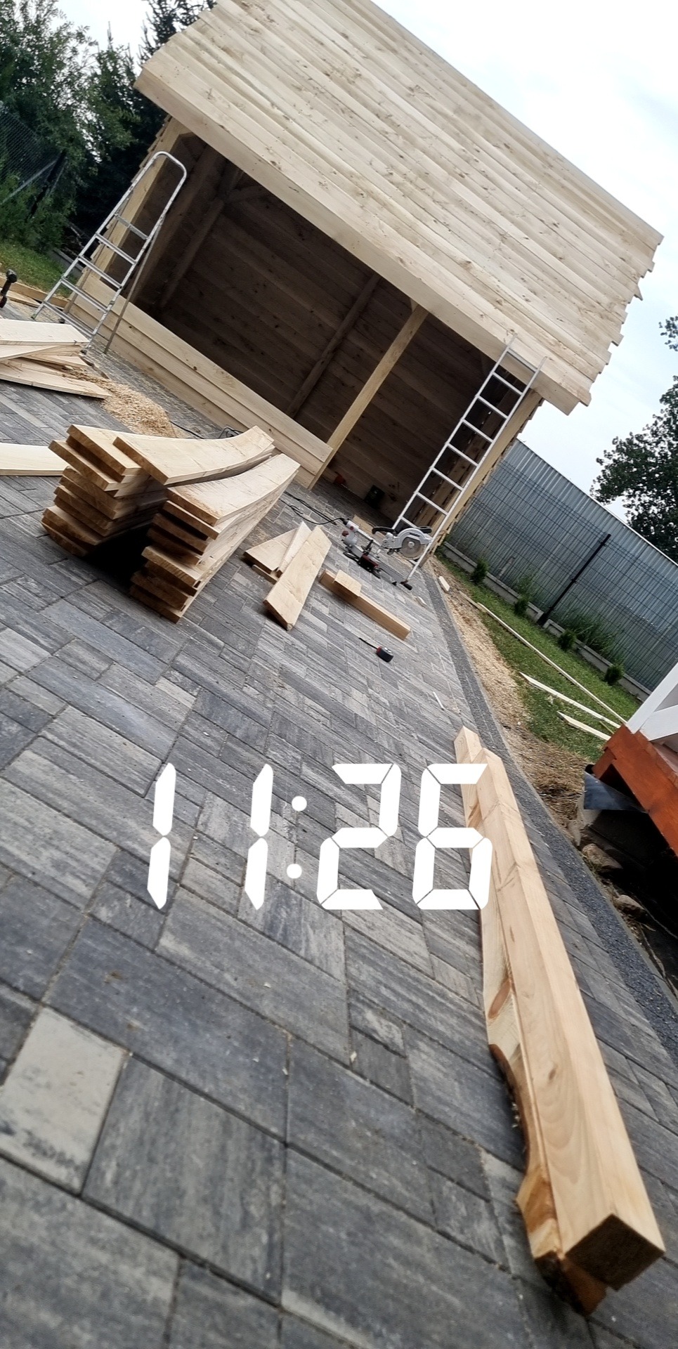 Drewniana konstrukcja dachu w trakcie budowy, widoczne deski, drabina i narzędzia na kostce brukowej, z cyfrowym zegarem nałożonym na zdjęcie.
