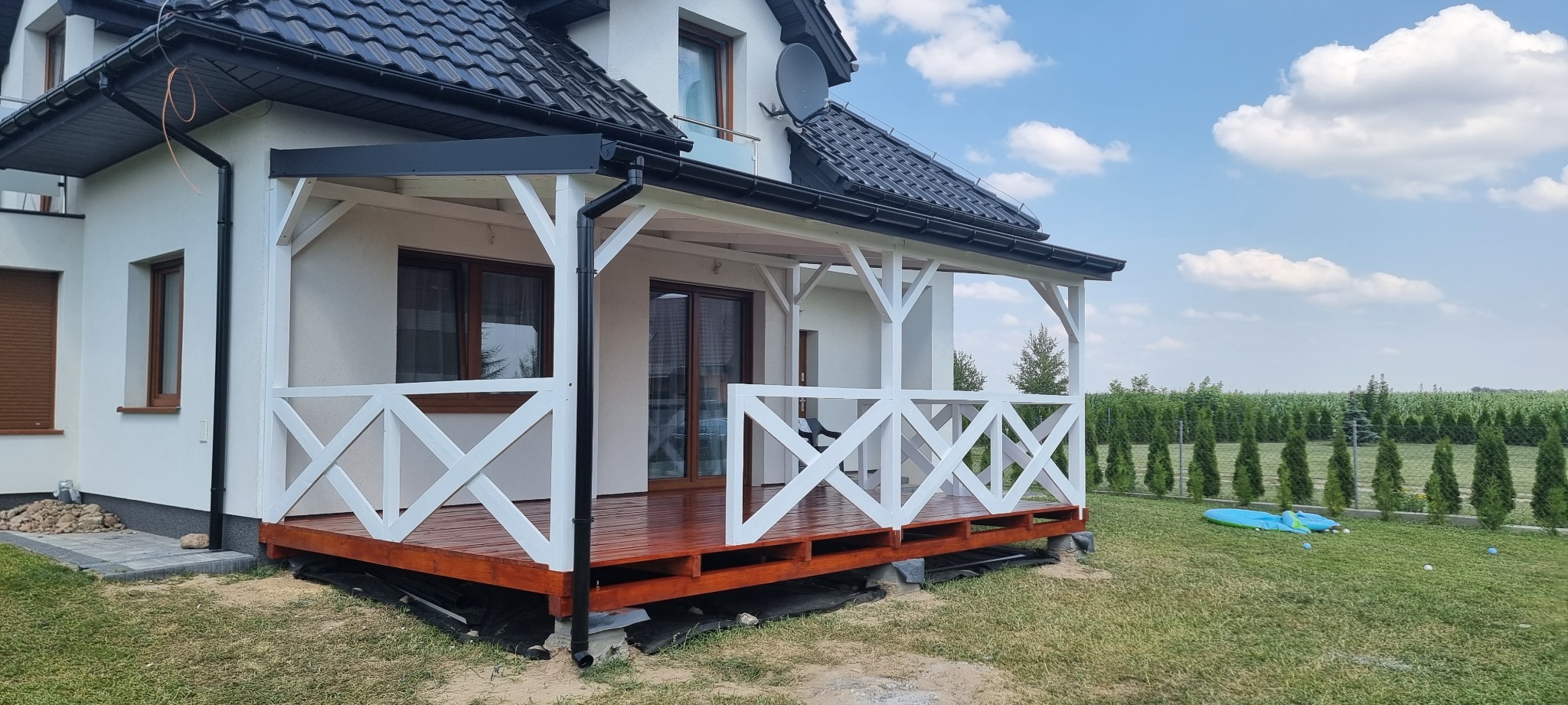 Nowoczesny dom z białymi ścianami i czarnym dachem, z dobudowanym drewnianym tarasem z białą balustradą w kształcie krzyży i brązową podłogą, widok na ogród z zielonym trawnikiem i rzędem tuj.