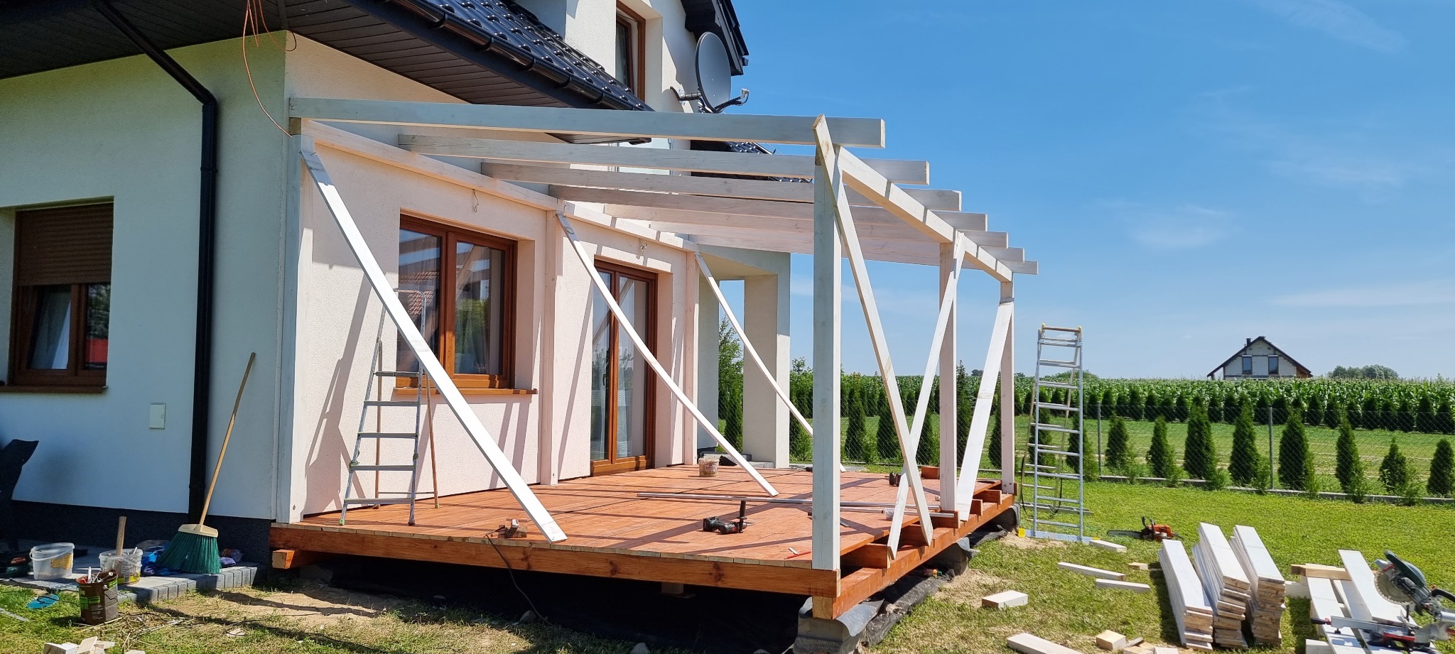 Drewniana konstrukcja pergoli w trakcie budowy przy ścianie domu jednorodzinnego, widoczne narzędzia i materiały budowlane, w tle zielony trawnik i rząd tuj.
