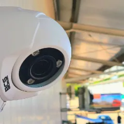 Kamera monitoringu BCS zamontowana na białej ścianie budynku przemysłowego, z widocznym fragmentem parkingu i ciężarówką w tle.