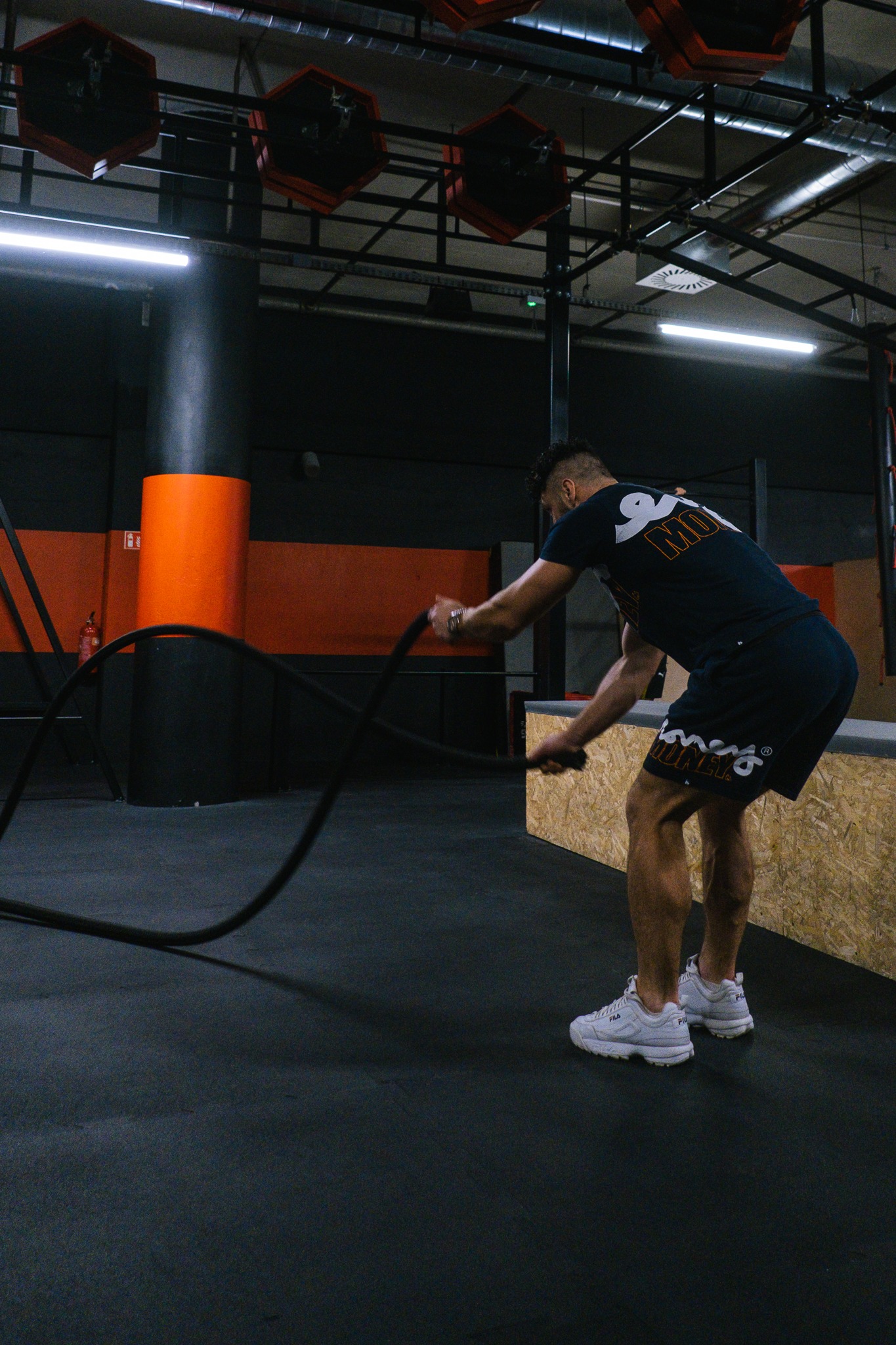 Mężczyzna w sportowym stroju ćwiczy z grubymi linami w siłowni, widoczne elementy konstrukcji sufitu i pomarańczowy słup.