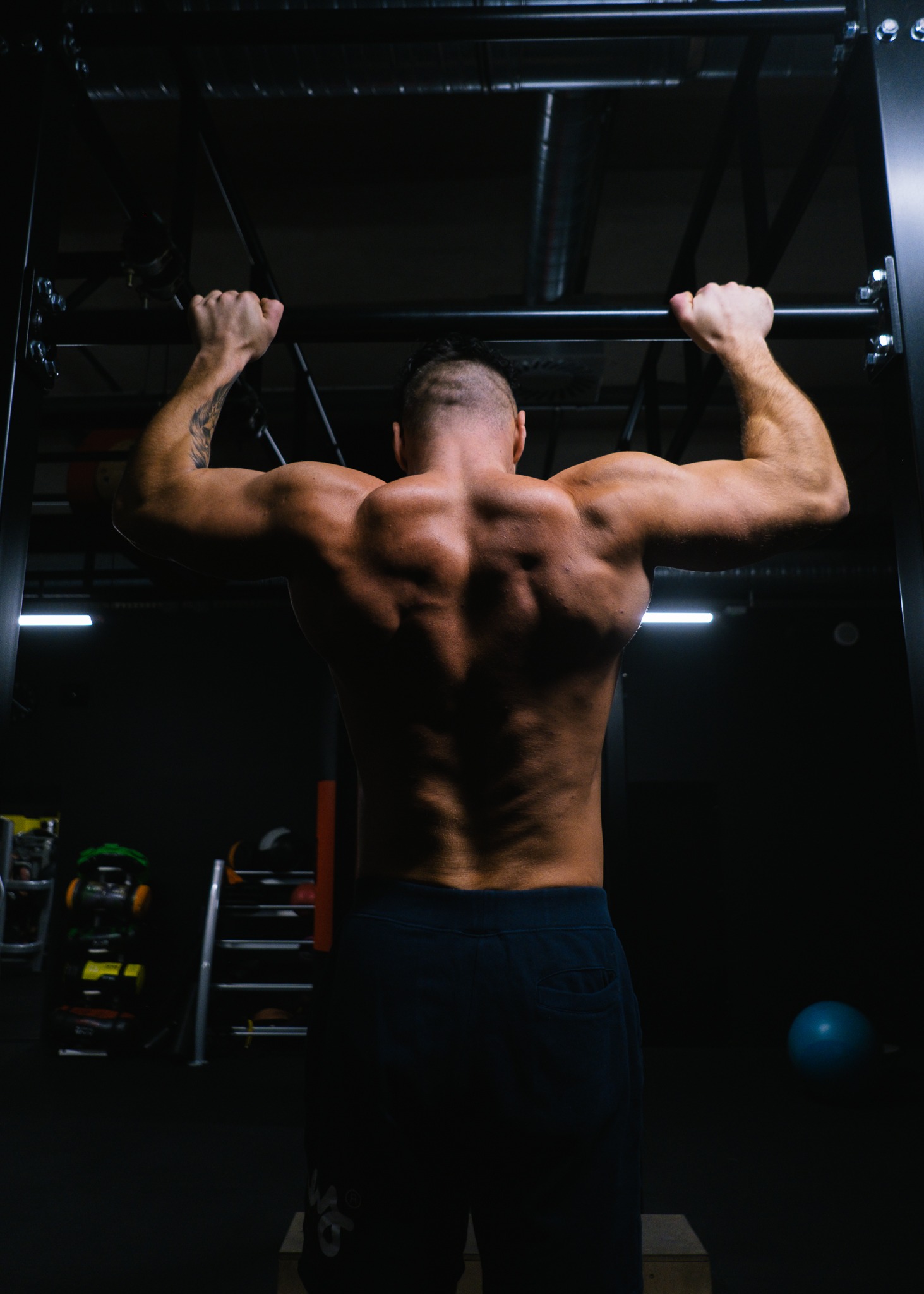 Mężczyzna o wyrzeźbionych plecach wykonuje podciąganie na drążku w siłowni, widok z tyłu, tatuaż na ramieniu.
