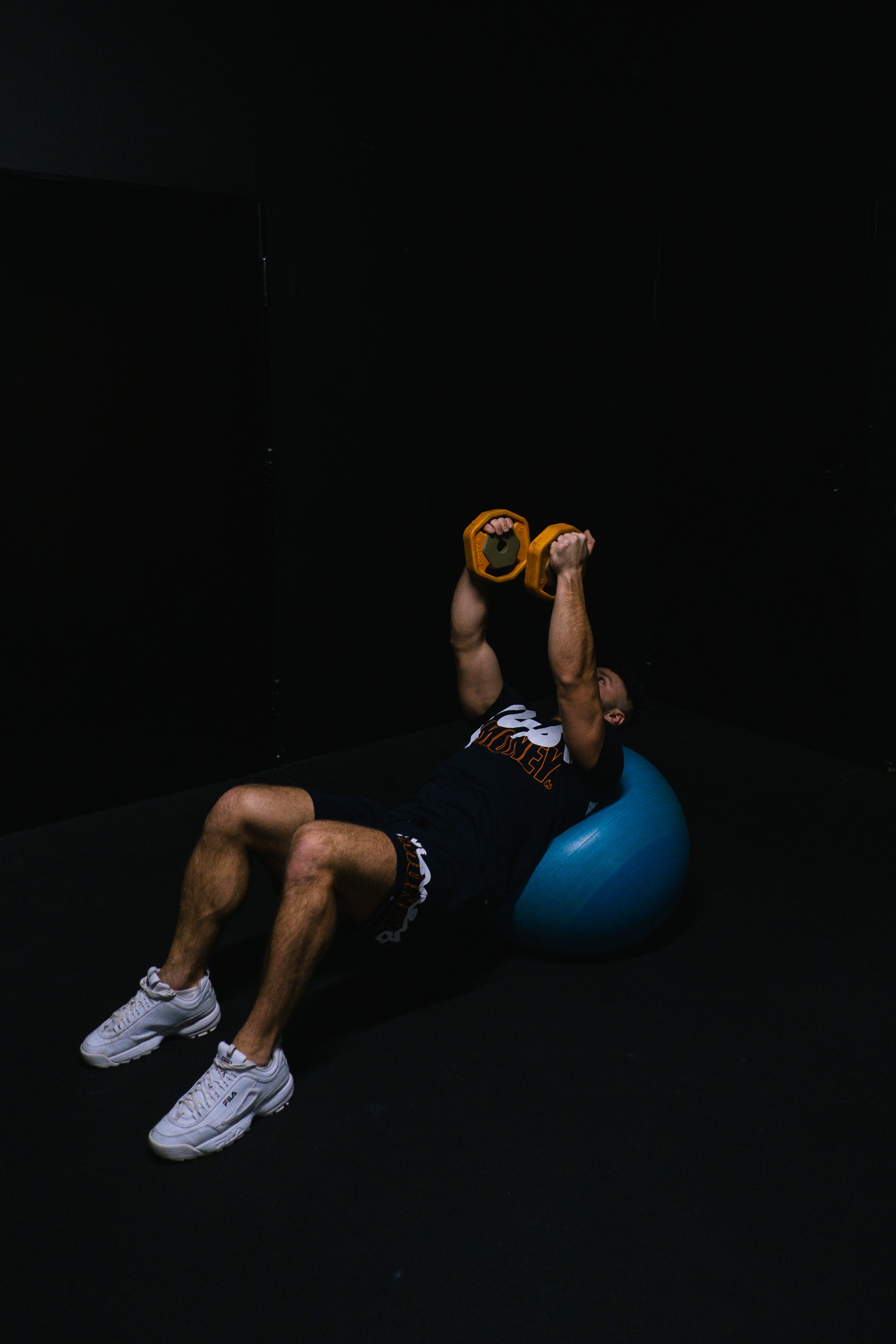 Mężczyzna w Mogilanach ćwiczy na piłce fitness z żółtymi obciążeniami w dłoniach, ubrany w sportowe szorty i koszulkę, na ciemnym tle.