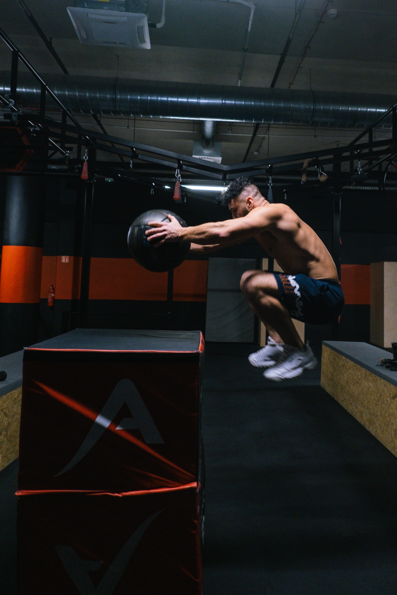 Mężczyzna bez koszulki, w krótkich spodenkach i białych butach, skacze z piłką lekarską nad skrzynią w siłowni.