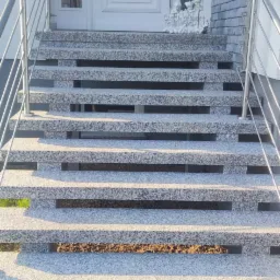 Zewnętrzne schody wejściowe z szarego kamienia z metalową balustradą, prowadzące do białych drzwi wejściowych z przeszklonym panelem bocznym i dekoracyjną kołatką, z widoczną dekoracją powitalną.