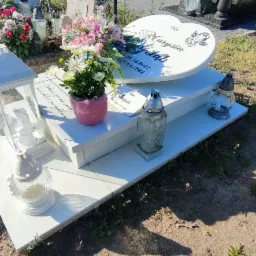 Biały nagrobek z sercem i napisem 'Marysia Gołąb' otoczony kwiatami i lampionami na cmentarzu, widok z góry na ziemię i trawę.