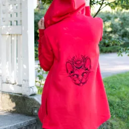Czerwona bluza z kapturem, z tyłu czarny haftowany wzór głowy kota z okiem w trójkącie, osoba na schodach w parku.