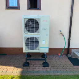 Zewnętrzna jednostka pompy ciepła Atlantic Inverter na specjalnej konstrukcji wsporczej, umieszczona na brukowanym patio obok ściany budynku z dwoma oknami i rynną spustową. Widoczny kran...