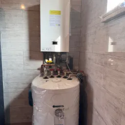 Biały piec gazowy Beretta i podgrzewacz wody Kospel SWK-120 w folii ochronnej, zamontowane na ścianie wyłożonej płytkami imitującymi drewno.