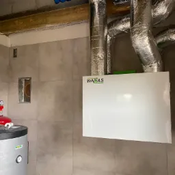 Biała jednostka centralna rekuperacji WANAS z podłączonymi, zaizolowanymi przewodami wentylacyjnymi w pomieszczeniu z szarą glazurą, widoczny bojler z czerwonym osprzętem.