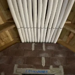 Równolegle ułożone, białe, karbowane rury wentylacyjne przymocowane metalowymi obejmami do drewnianej konstrukcji dachu, widoczne nad murowanym otworem w ścianie z brązowych bloczków.