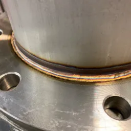 Szczegółowe zbliżenie spoiny kołnierzowej na rurze ze stali nierdzewnej, widoczne tęczowe przebarwienia termiczne.