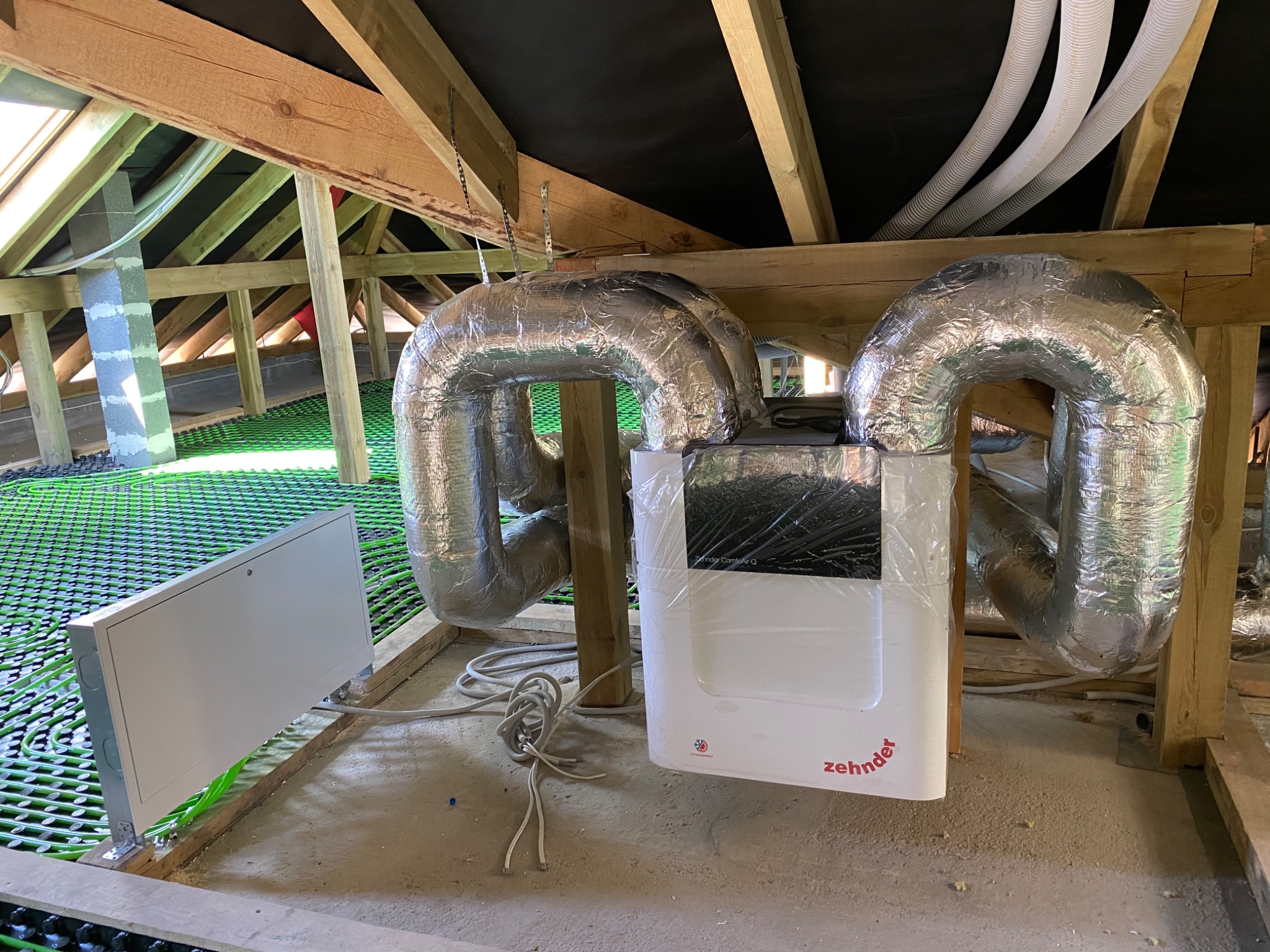 Centrala wentylacyjna Zhender ComfoAir Q w folii ochronnej, podłączona do izolowanych przewodów wentylacyjnych, zamontowana na poddaszu z widoczną konstrukcją dachu i instalacją ogrzewania...