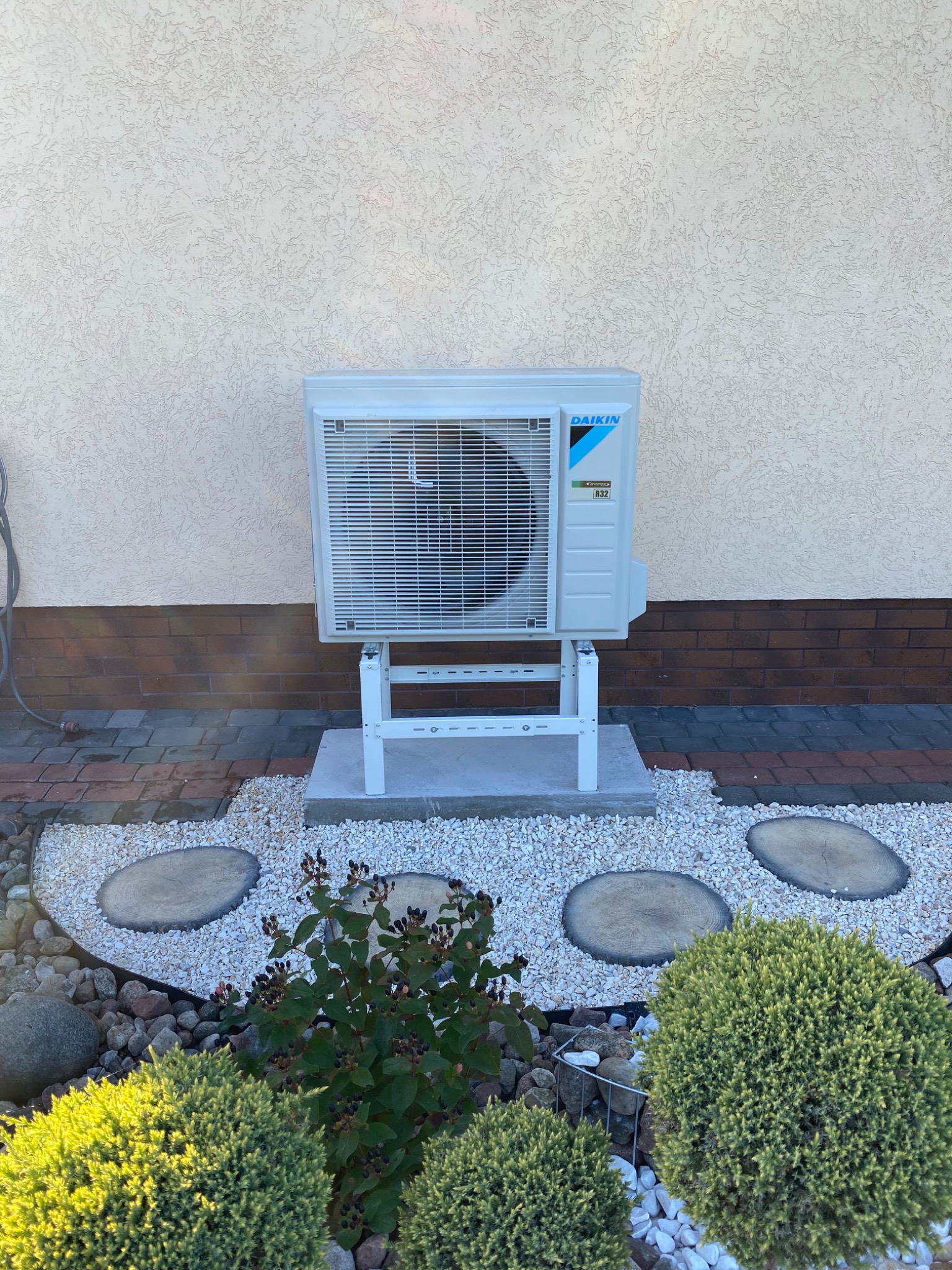 Zewnętrzna jednostka pompy ciepła Daikin na białym stelażu, ustawiona na betonowej podstawie, otoczona dekoracyjnym ogrodem z białym żwirem i drewnianymi krążkami