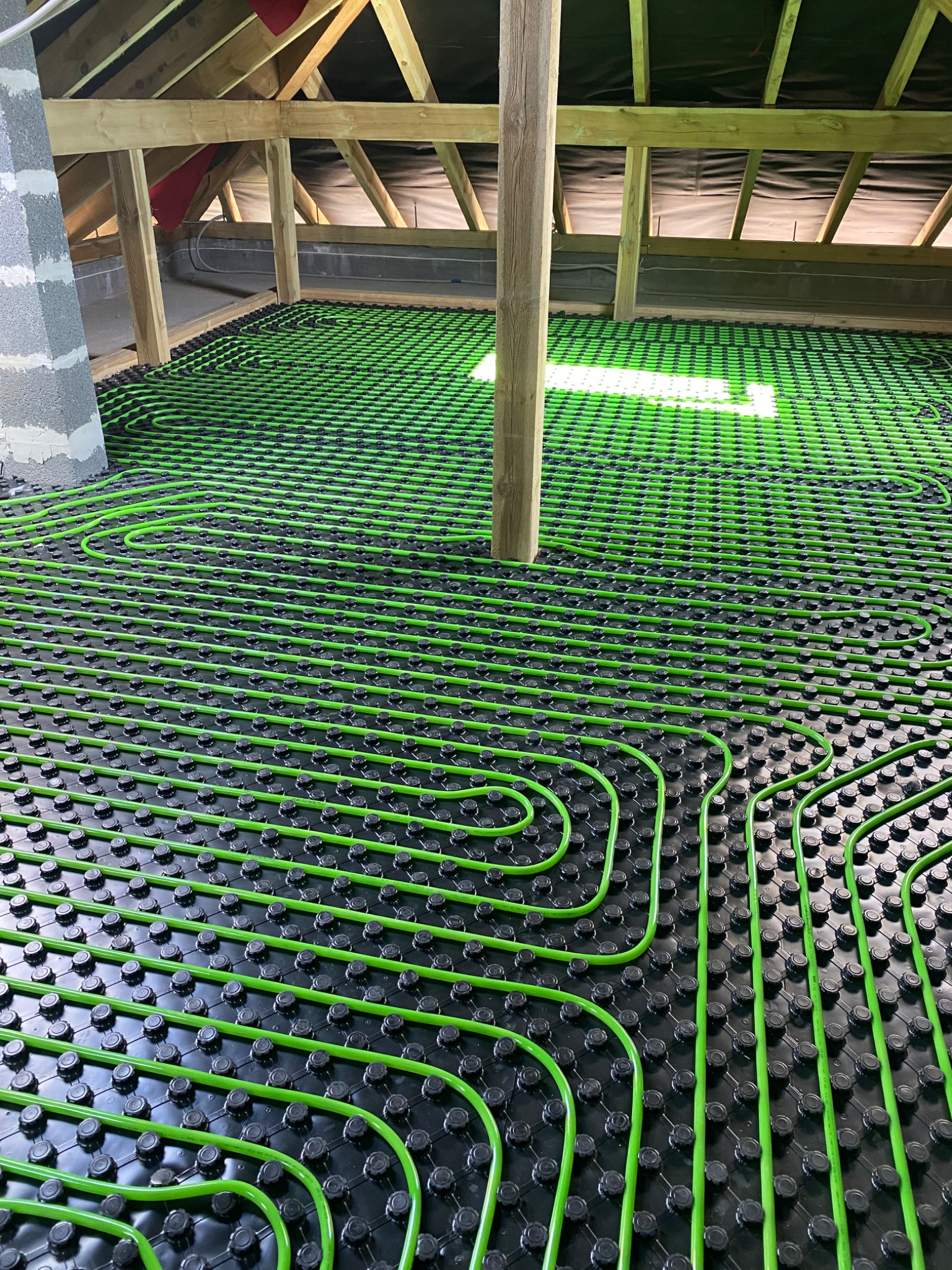 Zielone rury ogrzewania podłogowego ułożone na czarnej macie z mocowaniami, widok na poddasze z drewnianą konstrukcją dachu i słupem podtrzymującym.