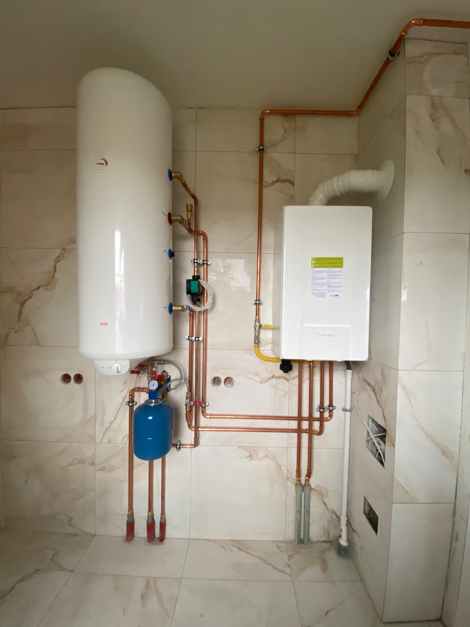 Zestaw kotła gazowego i zasobnika ciepłej wody użytkowej, połączonych miedzianymi rurami, zamontowany na ścianie wyłożonej marmurkowymi płytkami, z widocznym manometrem i zaworami.