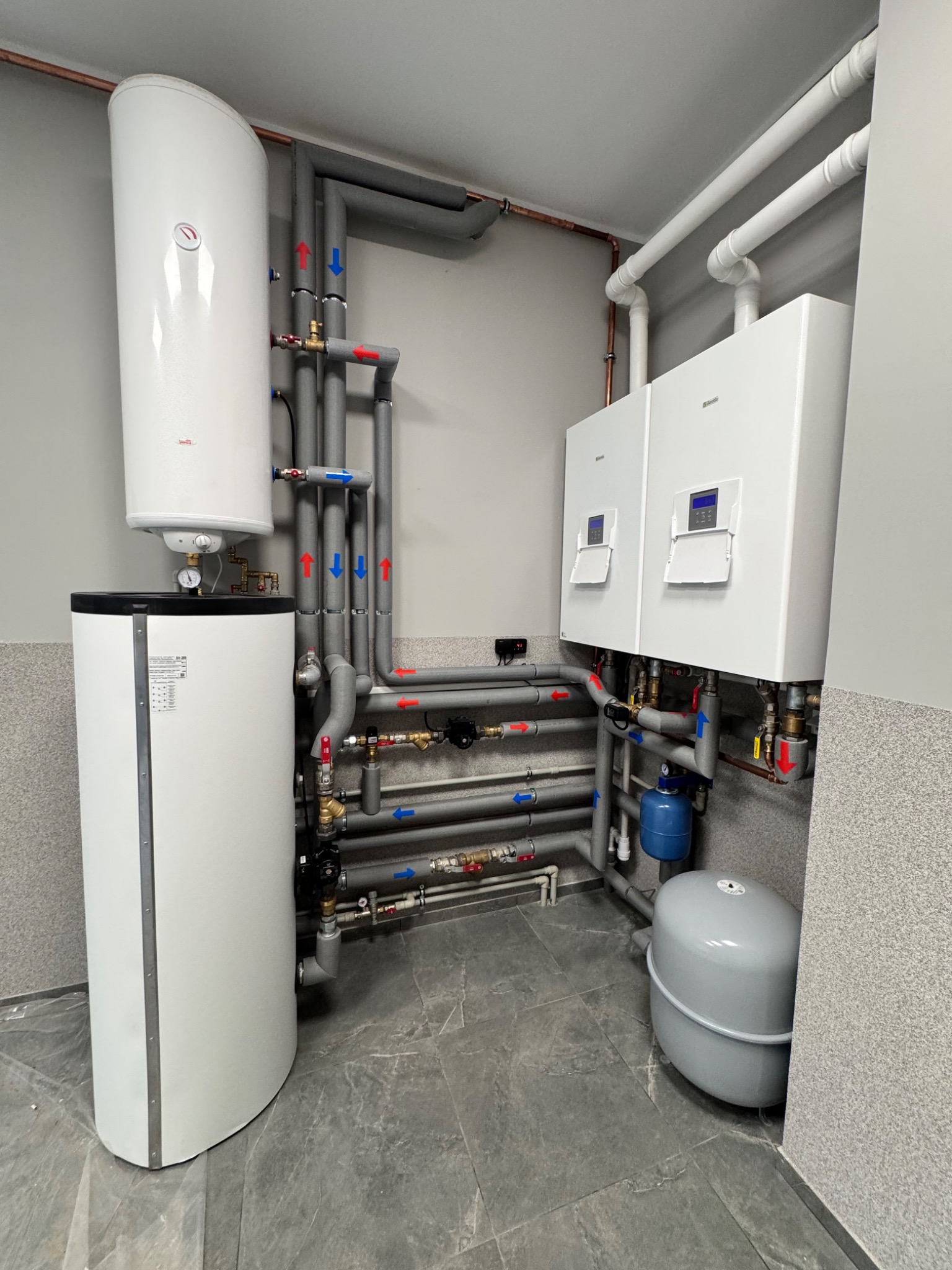 Nowoczesna kotłownia z dwoma piecami gazowymi Beretta, zasobnikiem ciepłej wody i szarymi rurami z oznaczeniami przepływu, na tle szarej ściany i podłogi z płytek.