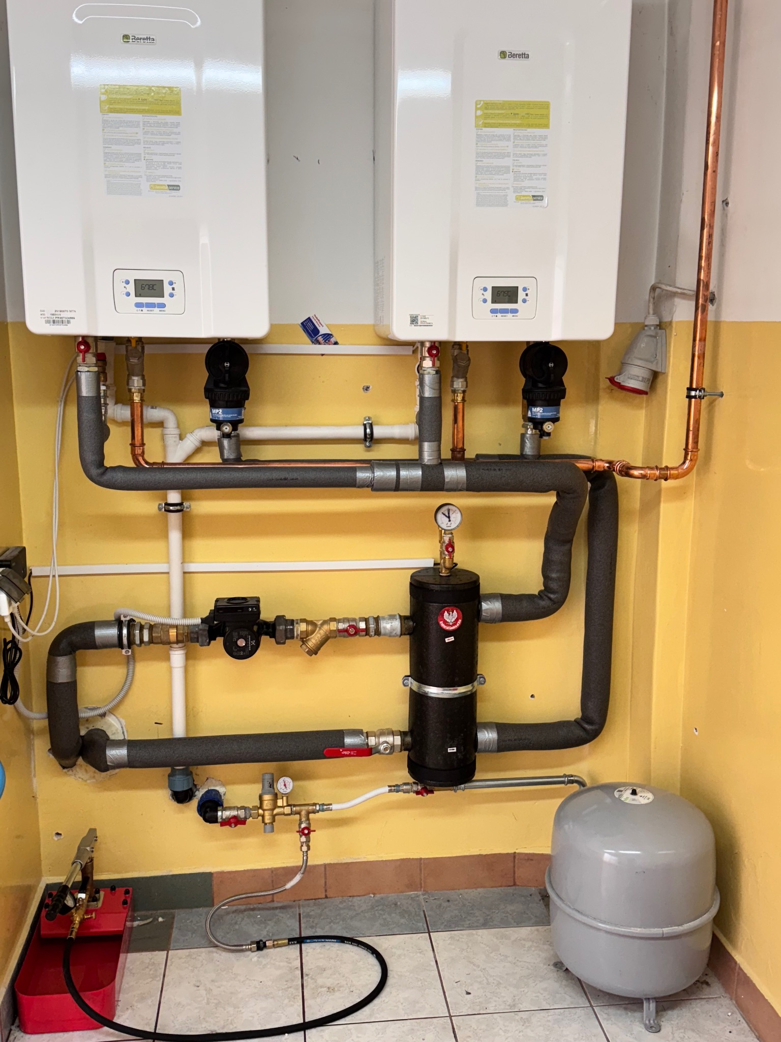 Dwa białe piece gazowe Beretta zamontowane na żółtej ścianie, z widoczną instalacją hydrauliczną i elementami kotłowni, w tym czarnym zbiornikiem i szarym naczyniem przeponowym.